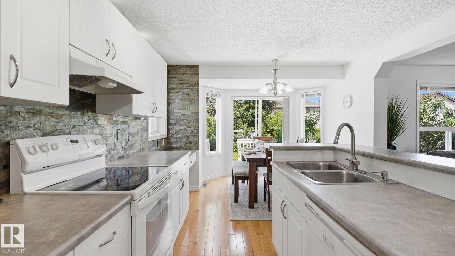 Kitchen featuring white cabinetry, stone-finish backsplash, wood-finish flooring, and a double basin sink - 3591 Mclean Crescent, Edmonton, AB