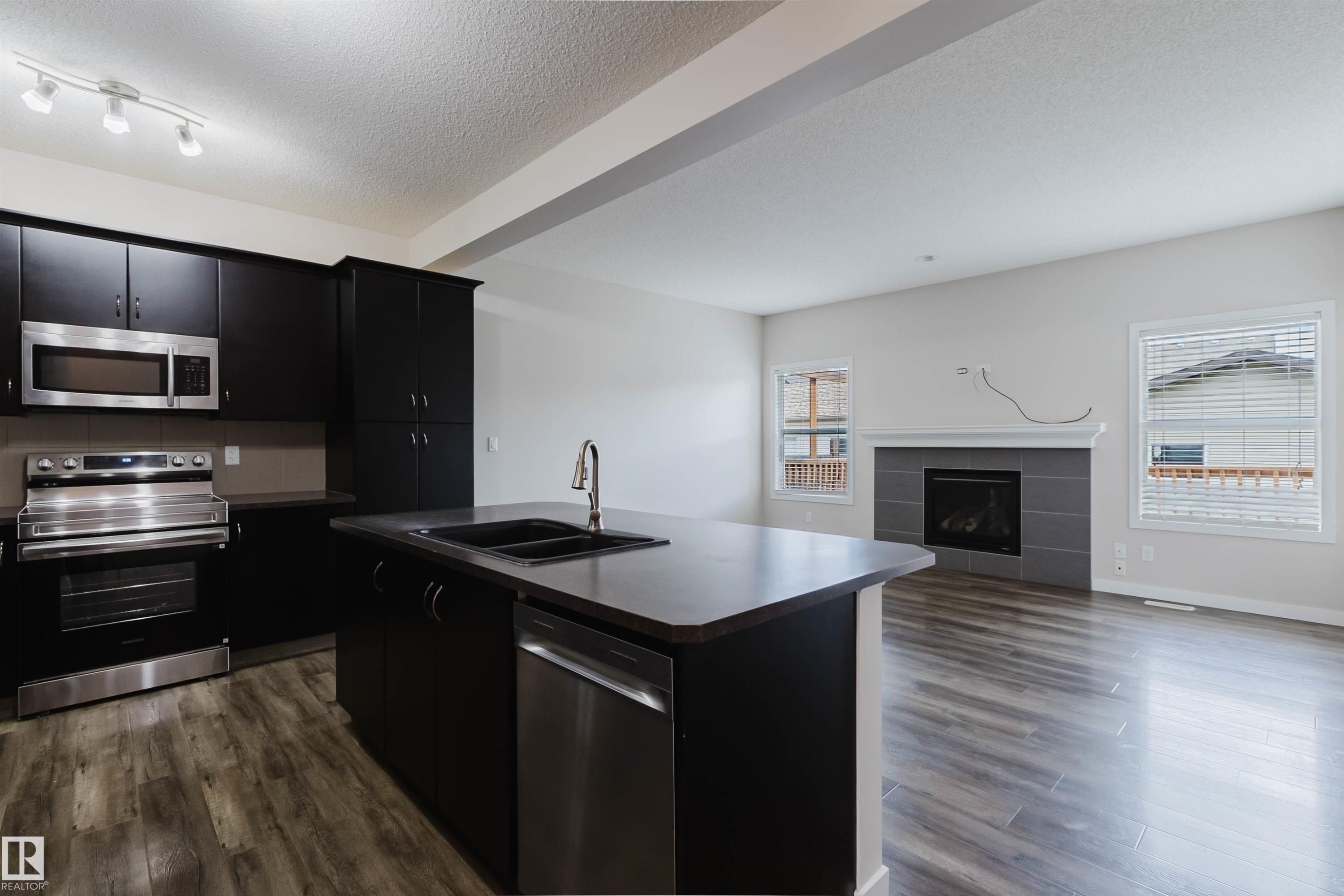 Open-concept kitchen and living area featuring dark cabinetry, stainless steel appliances, a kitchen island with a double basin sink, wood-finish flooring, and a fireplace with a tile surround - 636 Mcdonough Link Link, Edmonton, AB