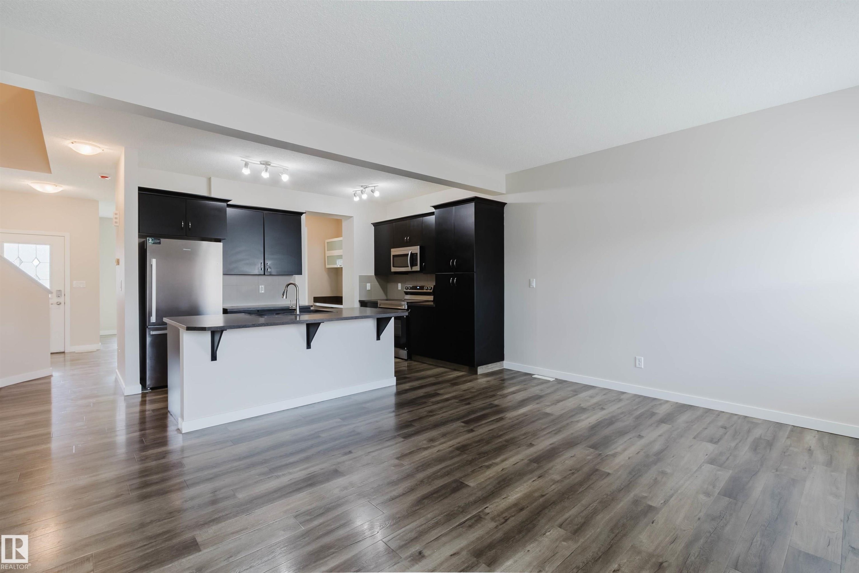 Open-concept living space featuring wood-finish flooring, a kitchen island with integrated sink, black cabinetry, stainless steel appliances, and track lighting - 636 Mcdonough Link Link, Edmonton, AB