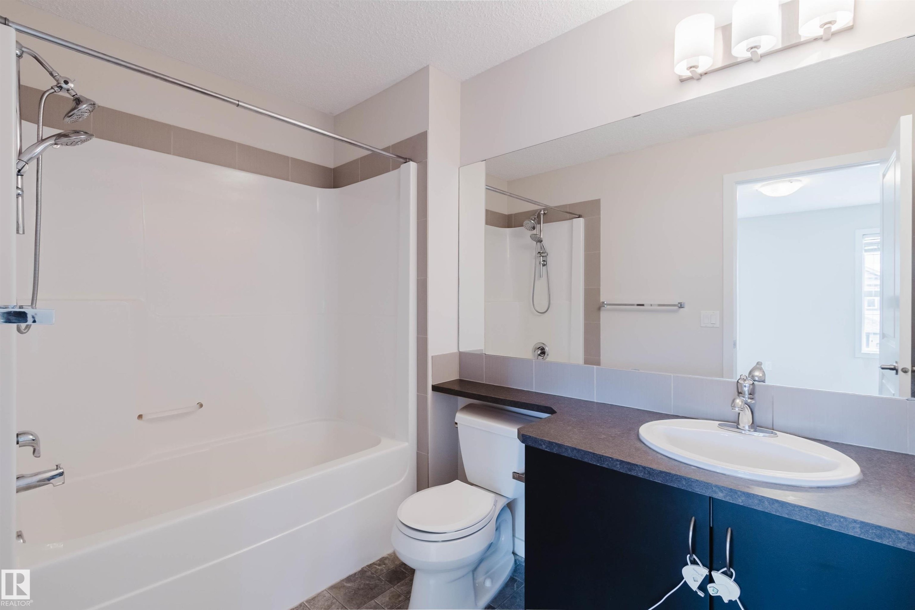 Bathroom featuring a tub/shower combination with a dual shower head, a single vanity with a dark countertop and undermount sink, a large wall-mounted mirror, and tile flooring - 636 Mcdonough Link Link, Edmonton, AB