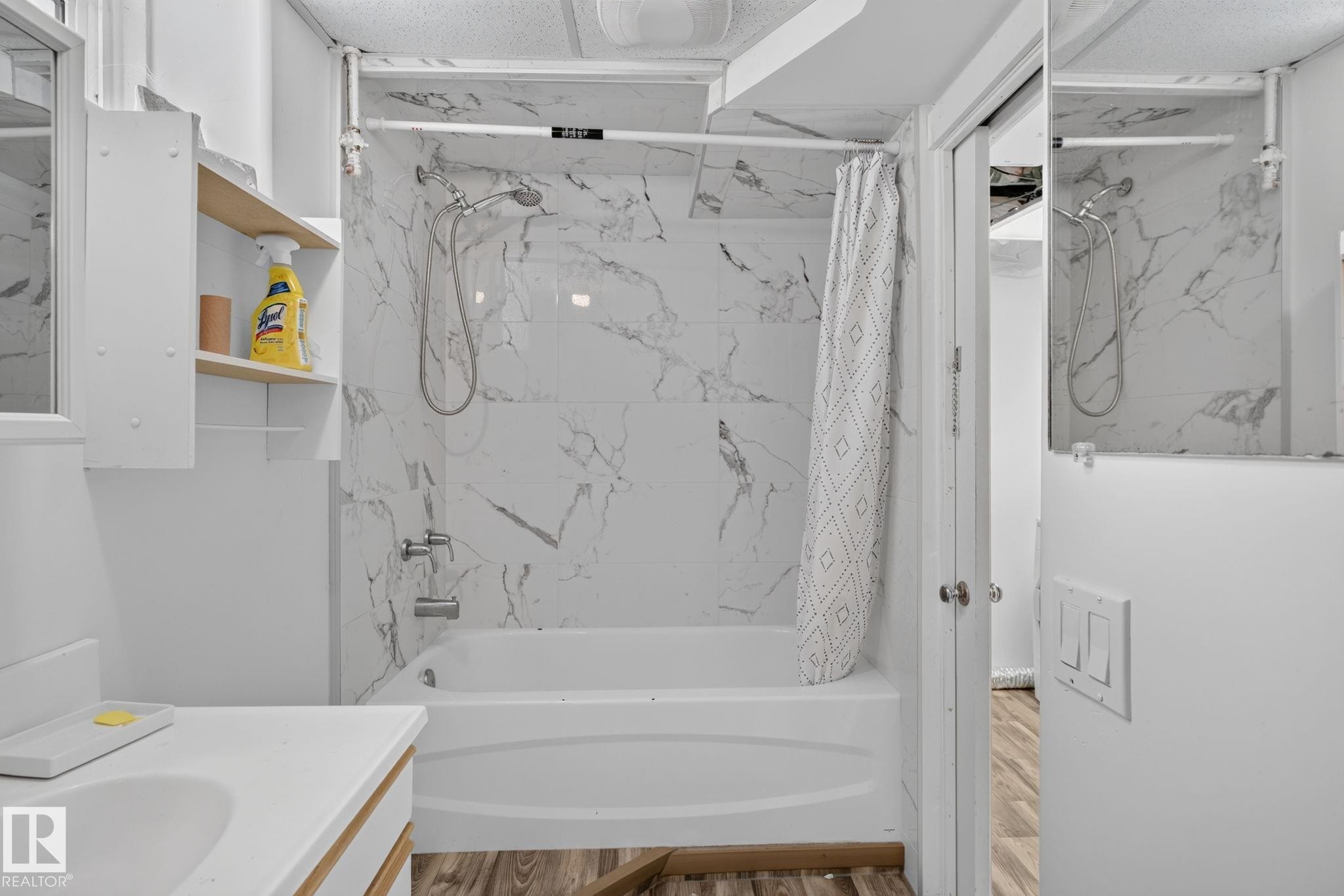 Bathtub with white marble-look surround and chrome fixtures, complemented by wood-finish flooring and a white vanity with an integrated sink - 12238 48 Street, Edmonton, AB