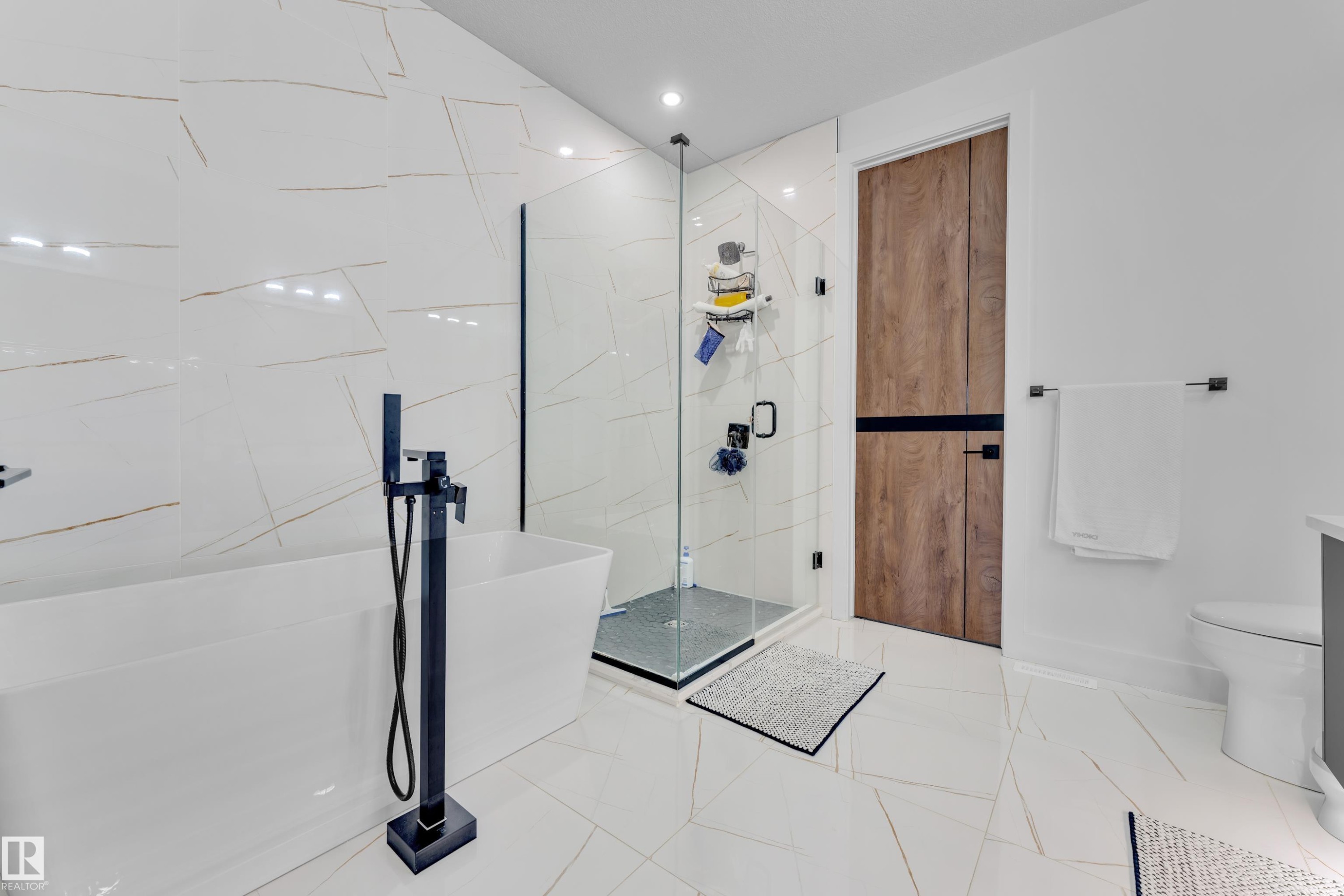 Contemporary bathroom featuring a freestanding soaking tub with a matte black floor-mounted faucet, a glass-enclosed shower with a dark framed door, and large format white tile flooring extending up the shower wall - 16816 49 Street, Edmonton, AB