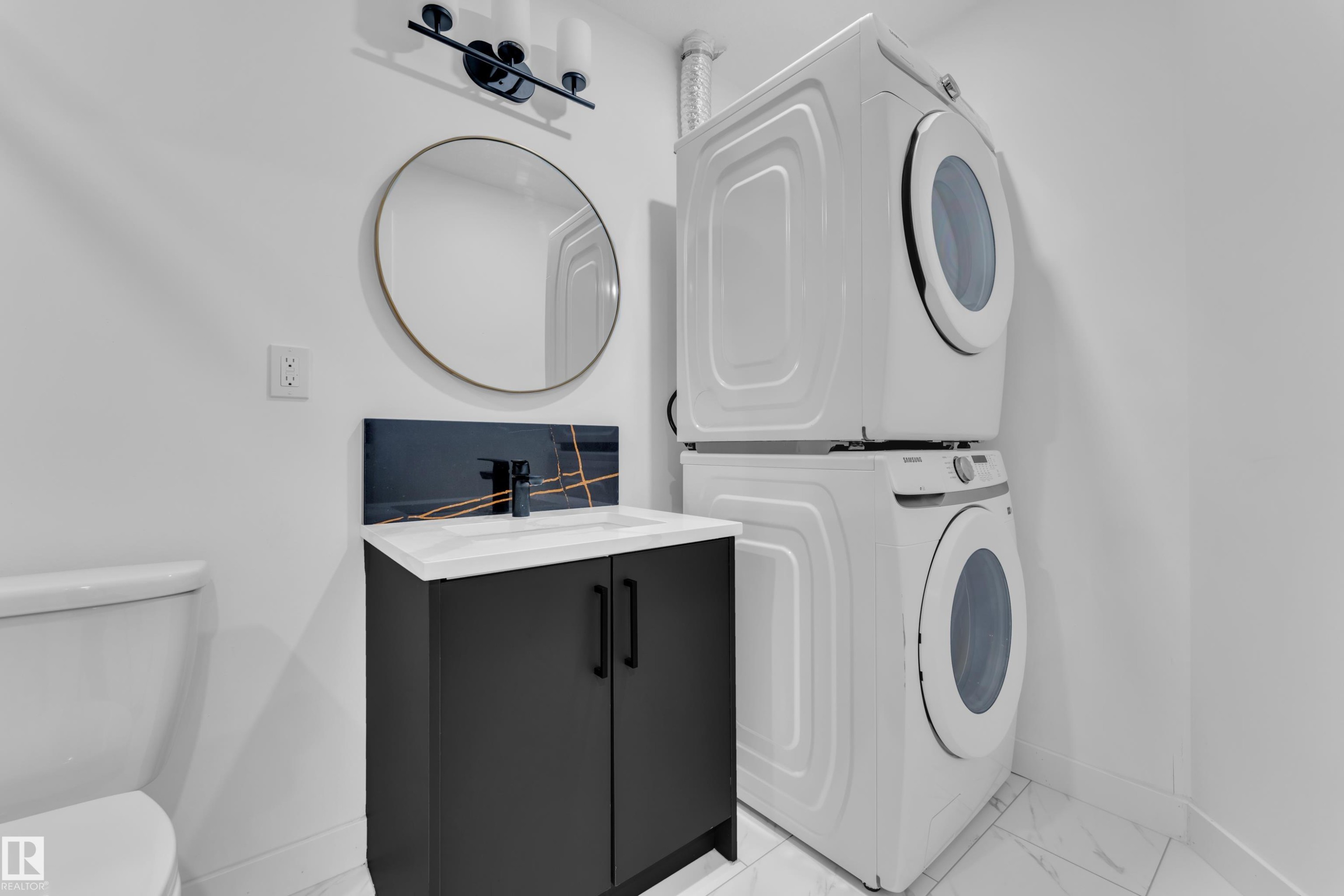 Compact laundry area featuring a stacked washer and dryer, a black vanity with white countertop, a round mirror, and patterned backsplash - 16816 49 Street, Edmonton, AB
