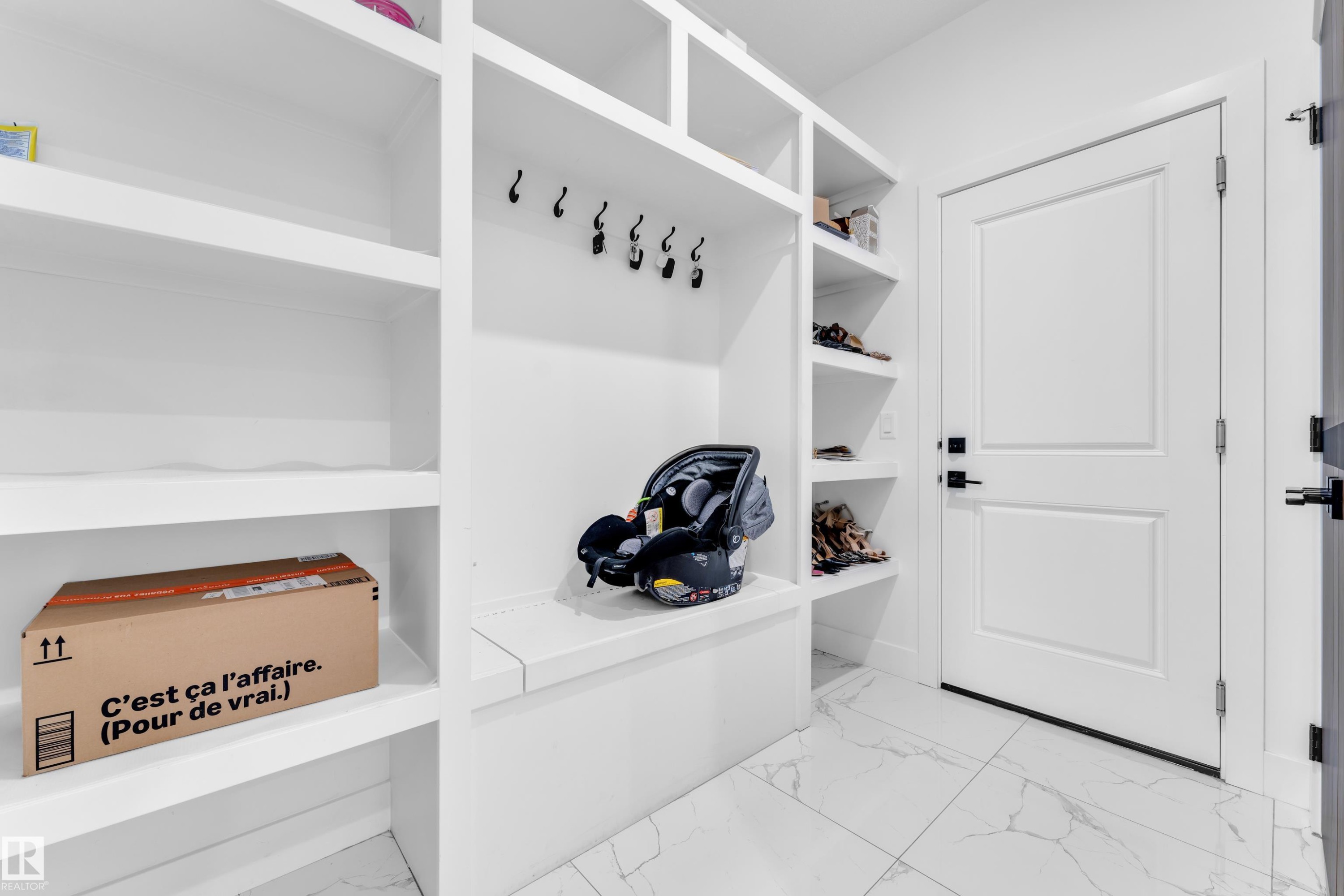 Built-in white shelving system with a bench, coat hooks, and additional shelving, complemented by white panel doors with matte black hardware, and marble-look tile flooring - 16816 49 Street, Edmonton, AB