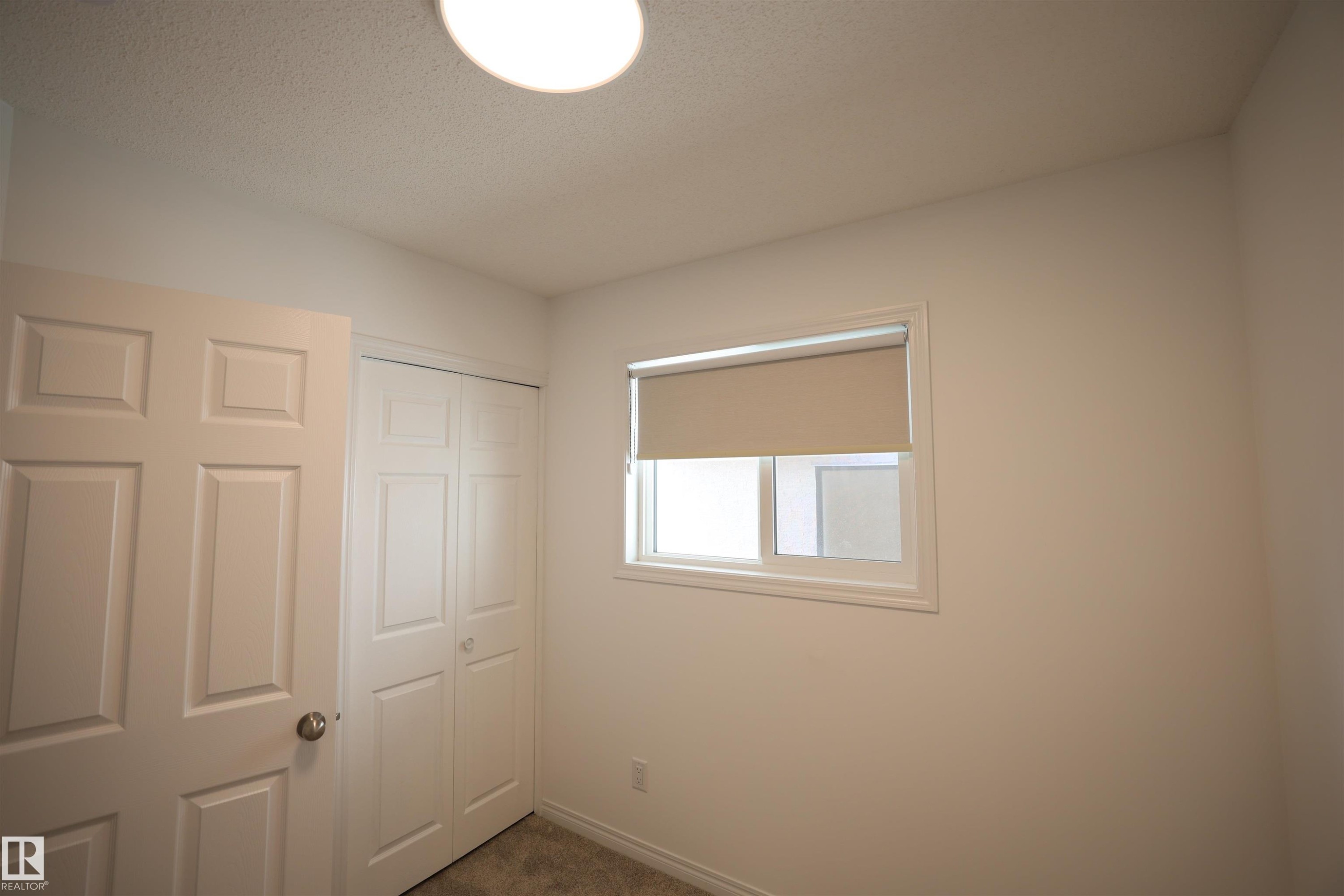 Room featuring white walls, a white paneled door, bi-fold closet doors, a white-framed window with a roller shade, and a flush-mount ceiling light fixture - 11120 132 Street, Edmonton, AB