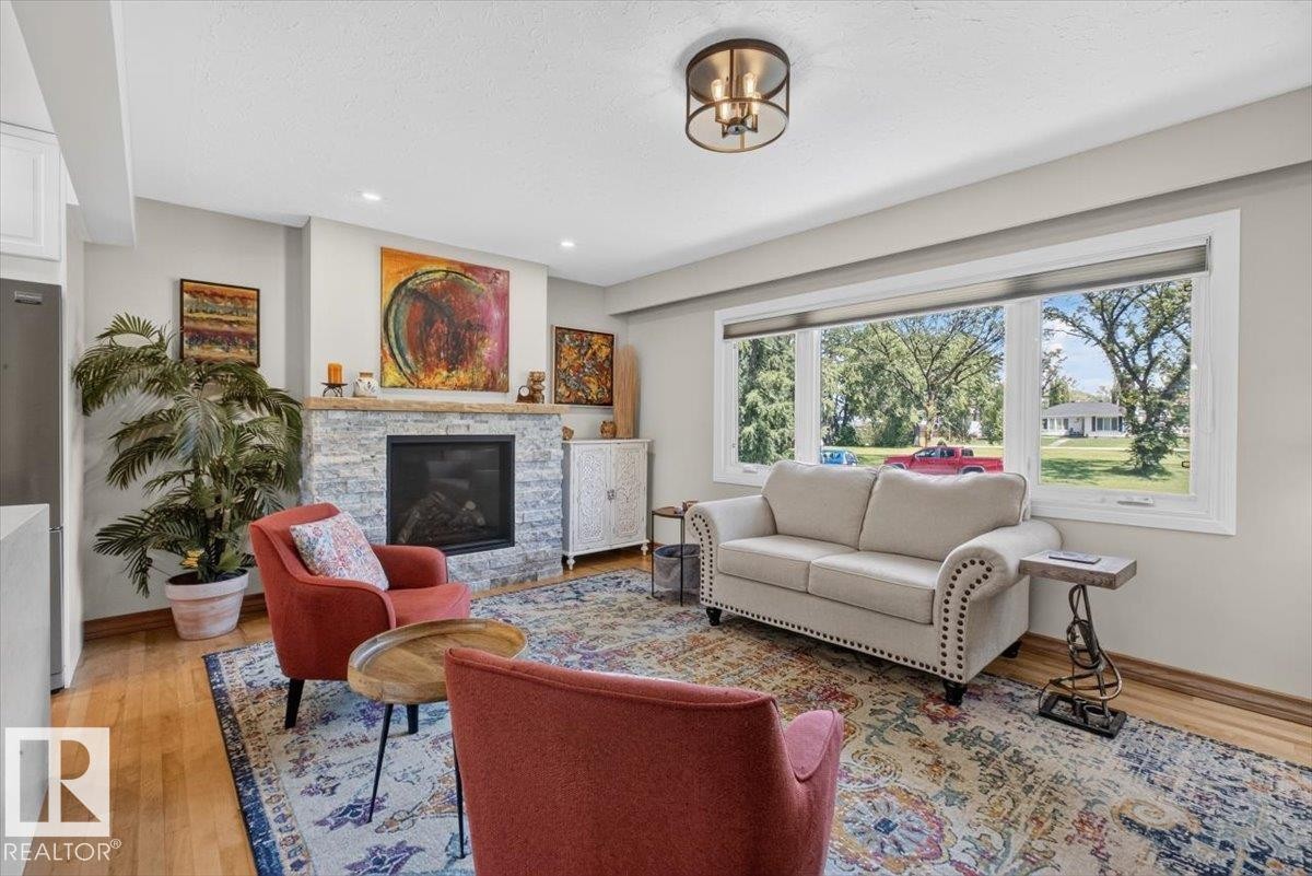 Living area featuring a stone-clad fireplace with a dark insert, a large picture window, wood-finish flooring, recessed lighting, and a contemporary ceiling fixture - 10736 48 Street Nw, Edmonton, AB - Indoor Photo Showing Living Room With Fireplace