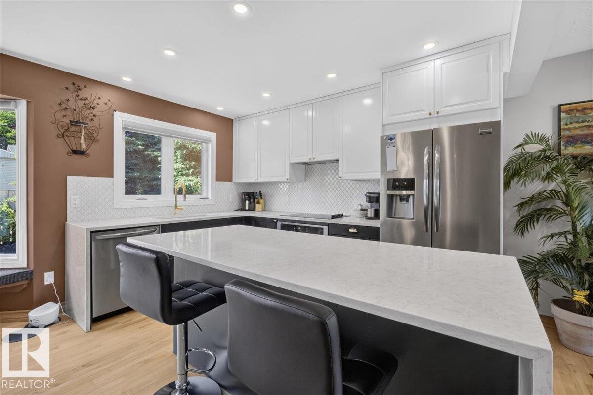 Kitchen featuring a large island with a light-toned countertop, white cabinetry, integrated stainless steel appliances, a white tile backsplash, and light wood-finish flooring - 10736 48 Street Nw, Edmonton, AB - Indoor Photo Showing Kitchen With Upgraded Kitchen