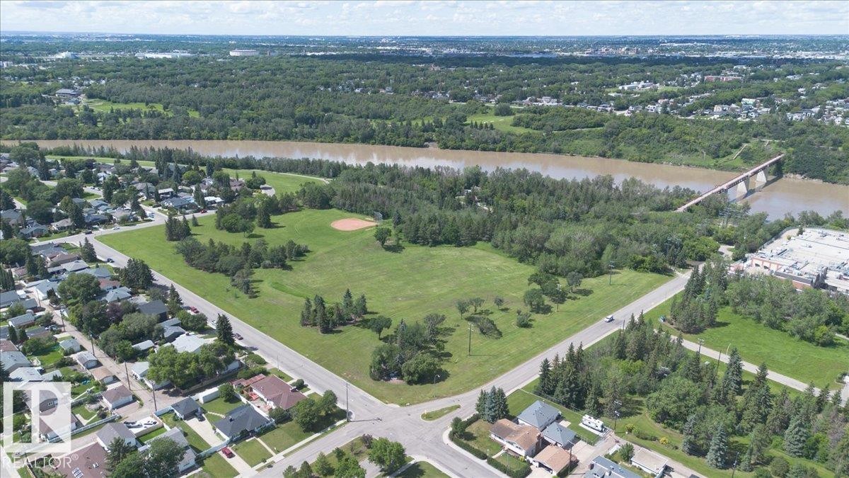 Aerial perspective showcasing a sprawling green space with a baseball diamond, surrounded by mature trees, residential properties, and a river with a bridge in the background - 10736 48 Street Nw, Edmonton, AB - Outdoor With View