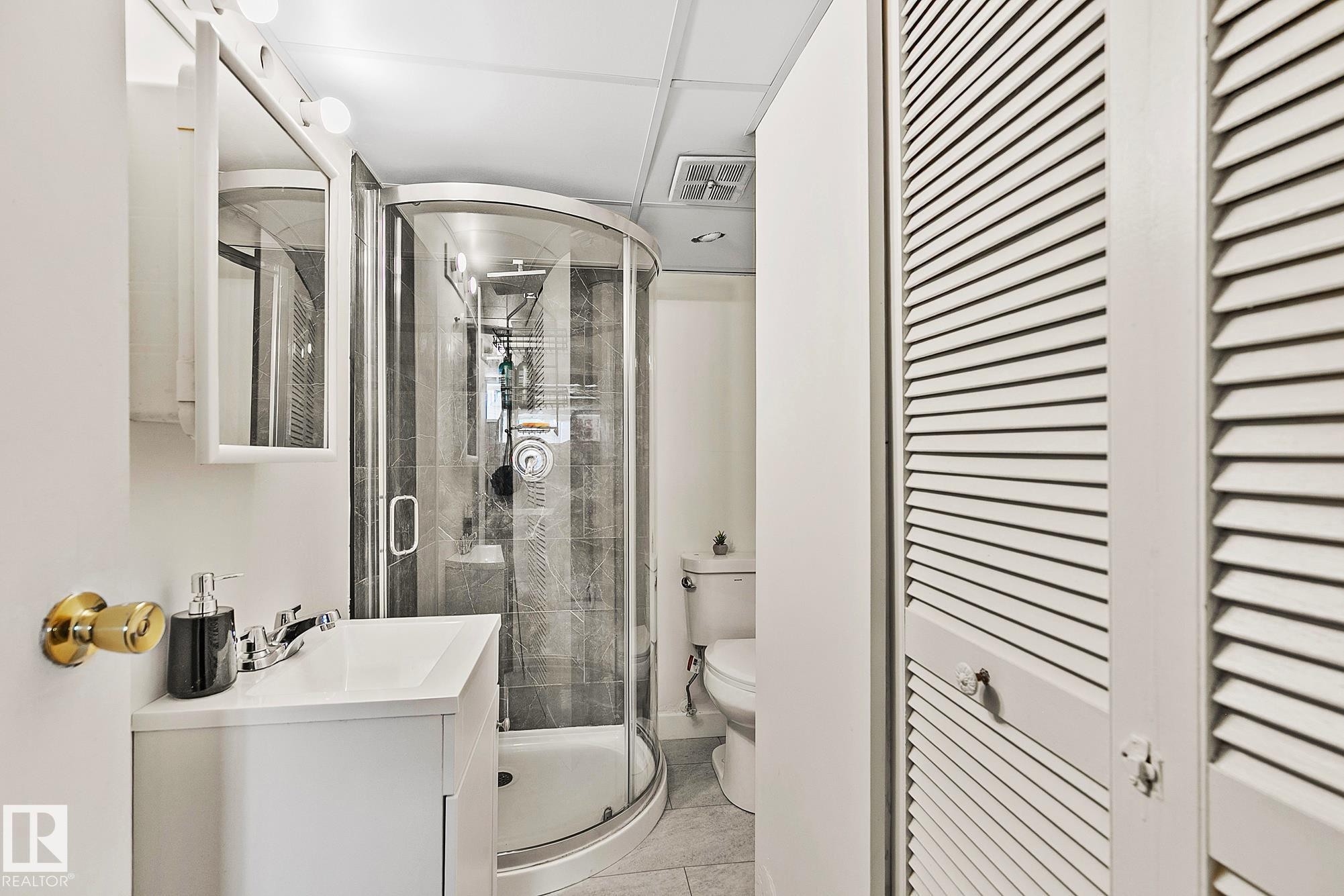 Bathroom featuring a curved glass shower enclosure, a white vanity with an integrated sink, a wall-mounted medicine cabinet, and louvered bi-fold doors - 17059 98 Street, Edmonton, AB - Indoor