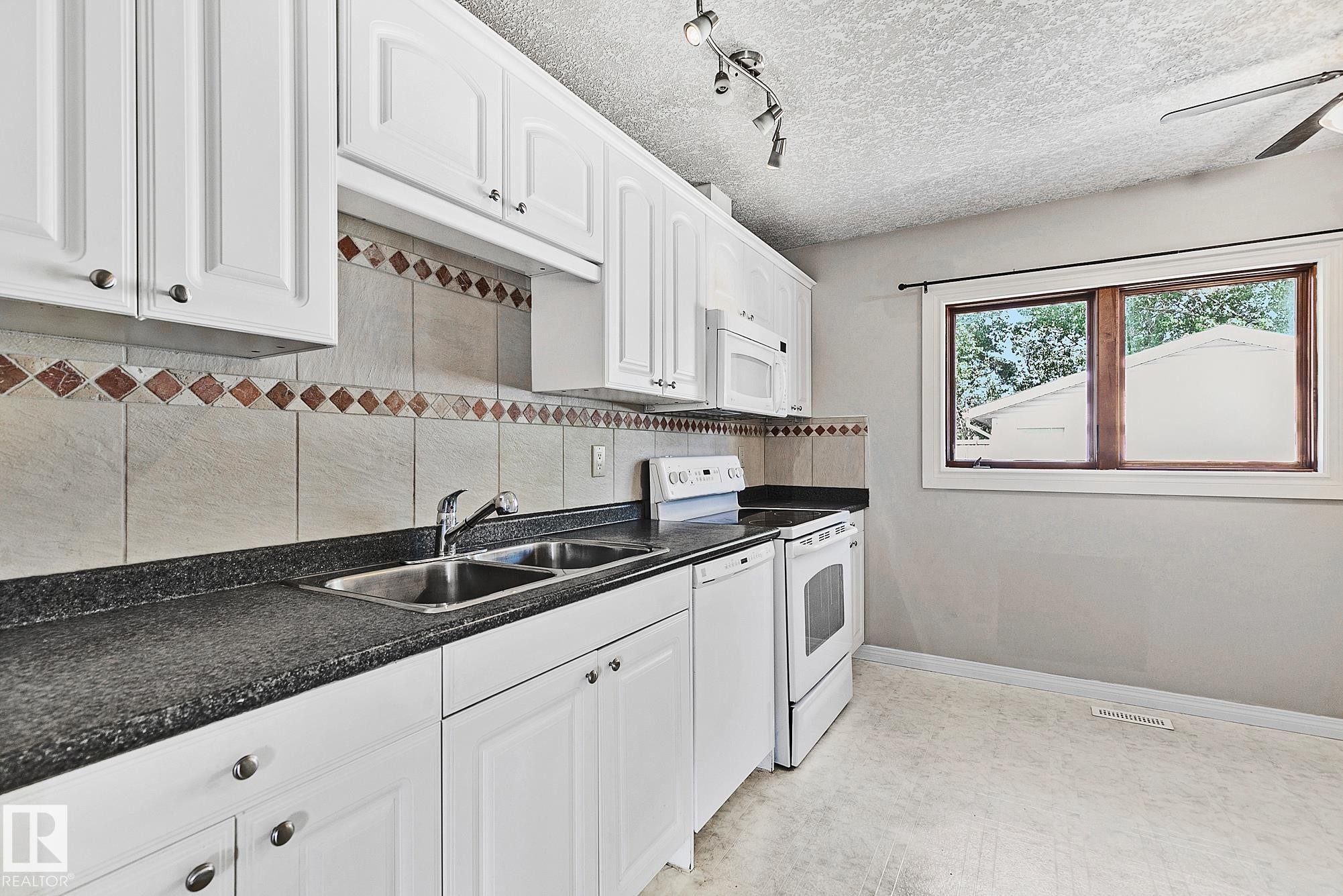 Kitchen featuring white cabinetry with brushed nickel hardware, speckled dark countertops, a double basin stainless steel sink, and a tile backsplash with decorative diamond accents - 17059 98 Street, Edmonton, AB - Indoor Photo Showing Kitchen With Double Sink