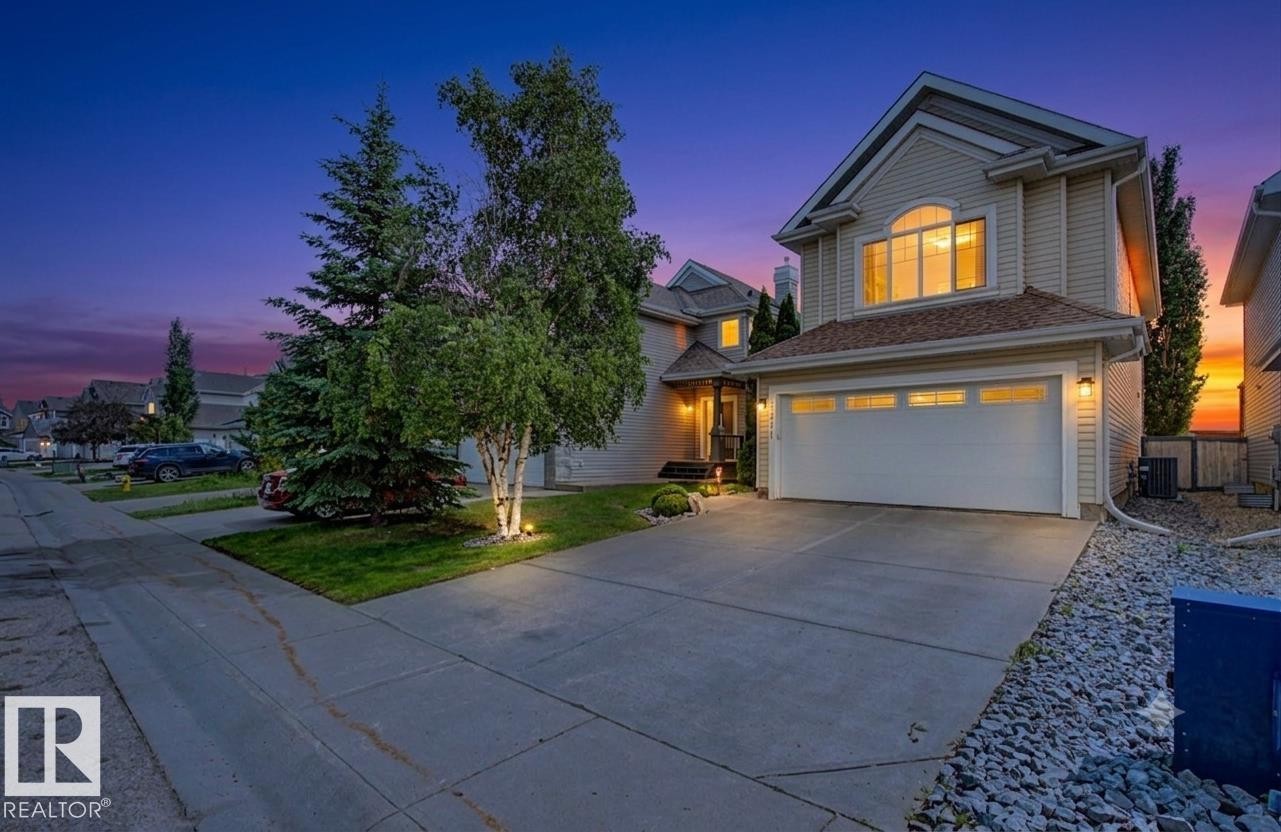 Two-story property featuring light-toned horizontal siding, an attached two-car garage with transom window accents, a concrete driveway, and a manicured front lawn with mature trees - 8311 11 Avenue Sw, Edmonton, AB