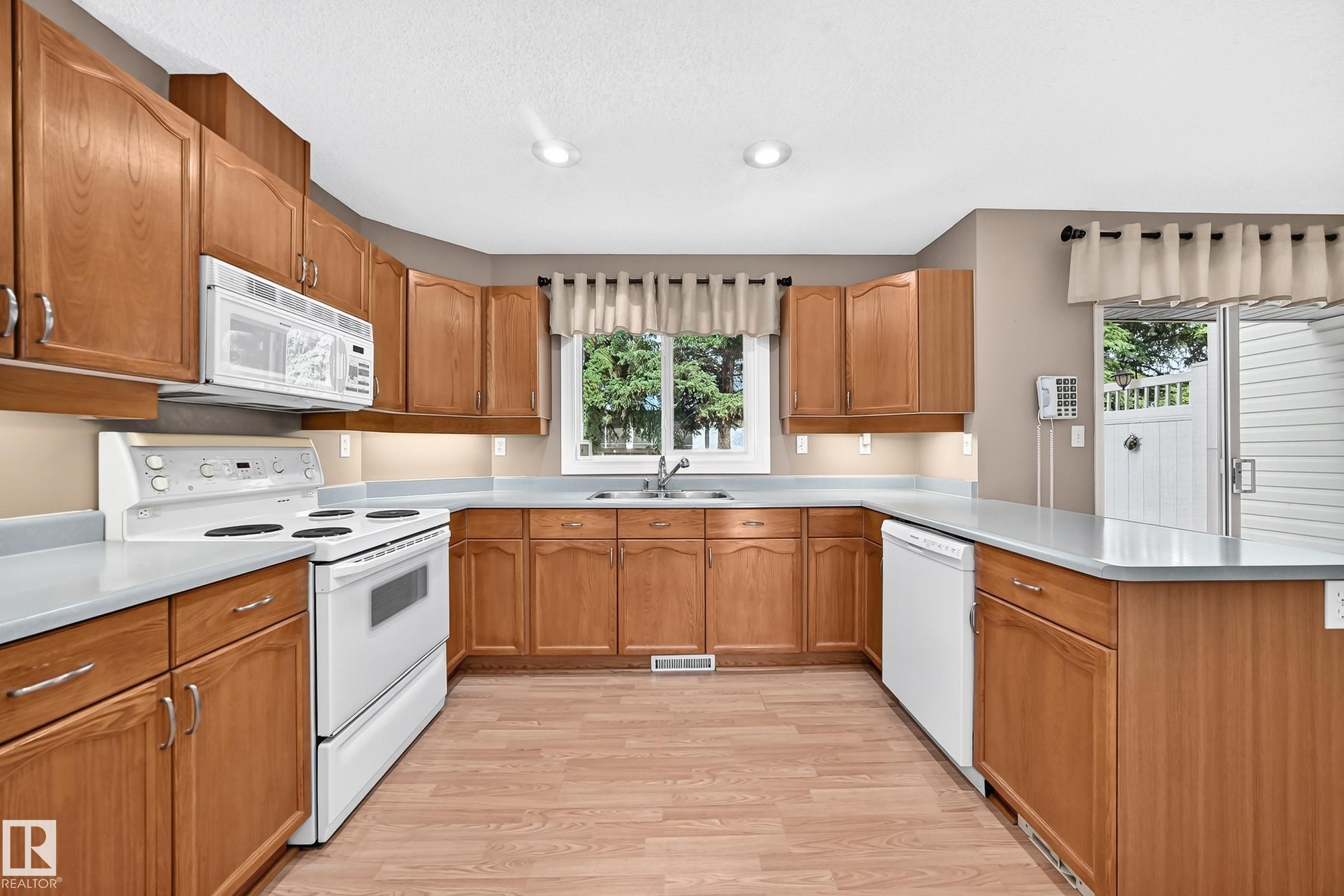 Bright kitchen featuring wood-finish cabinetry, white appliances, and light countertops - #51 9630 176 St Nw, Edmonton, AB - Indoor Photo Showing Kitchen With Double Sink
