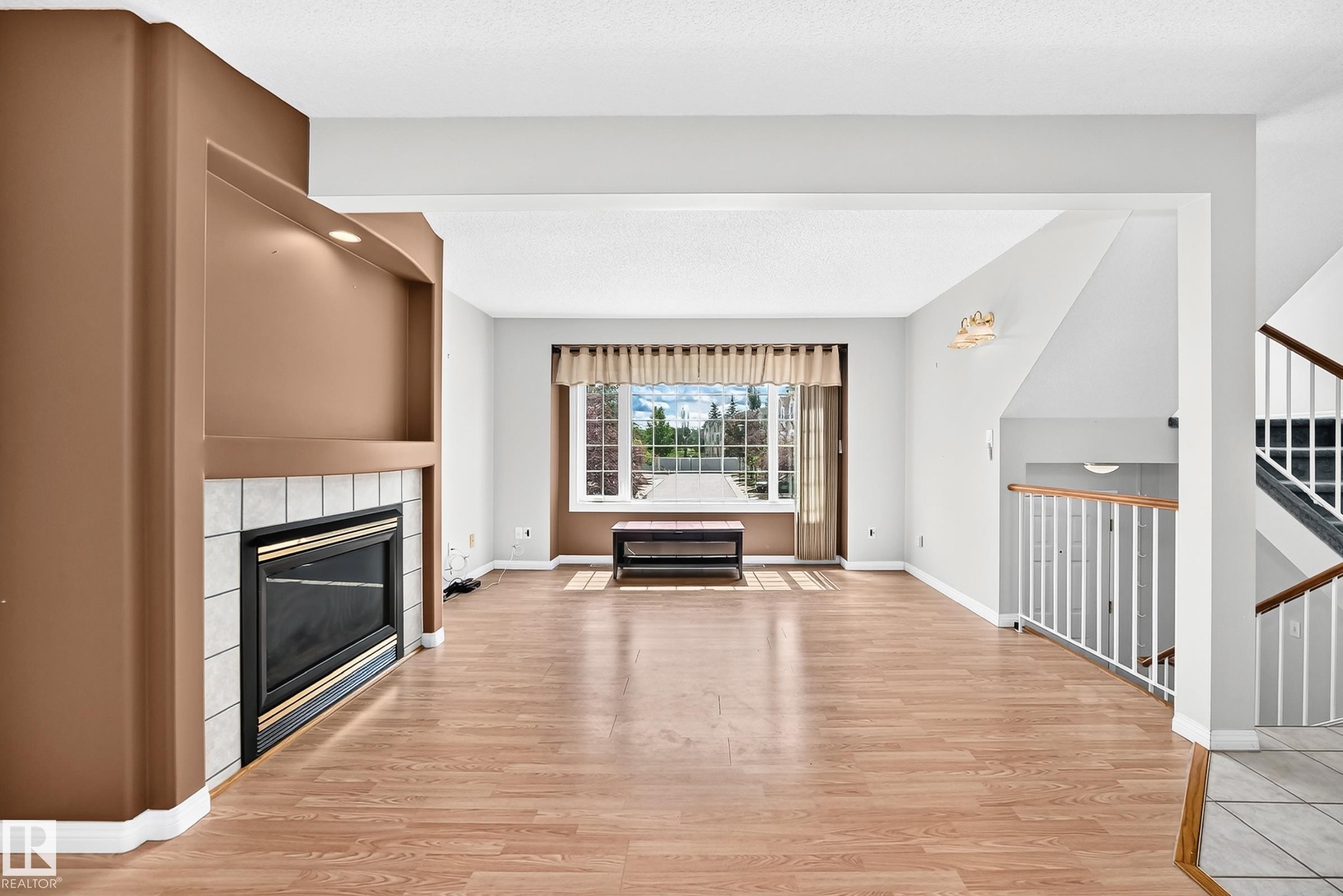 Living area featuring wood-finish flooring, a fireplace with a tiled surround, a large window with drapes, and a staircase with a white baluster and wood handrail - #51 9630 176 St Nw, Edmonton, AB - Indoor Photo Showing Living Room With Fireplace