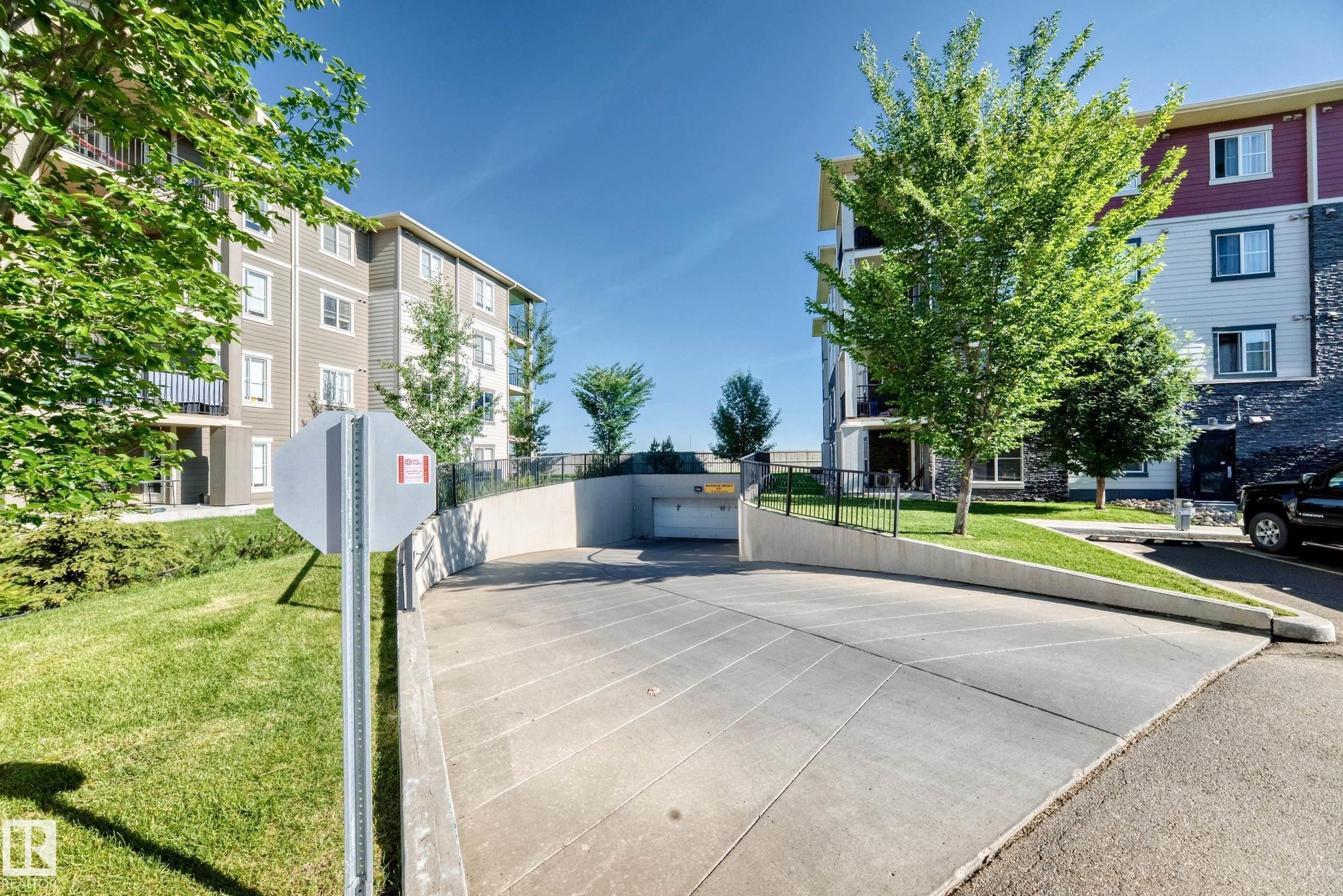 Concrete ramp leading to an underground parking garage, bordered by manicured lawns and mature trees - 308 18126 77 Street, Edmonton, AB