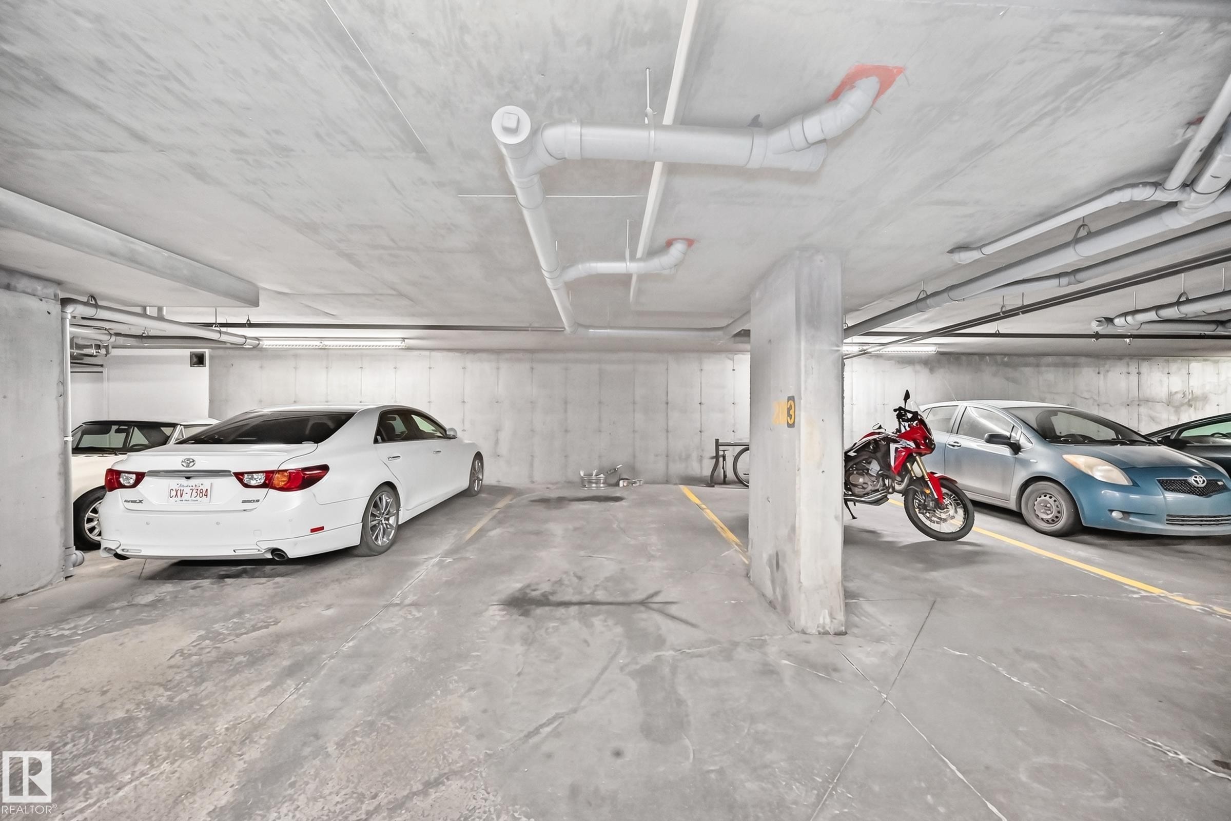 Concrete parking garage with exposed ceiling pipes, fluorescent strip lighting, and painted yellow parking lines - 308 18126 77 Street, Edmonton, AB