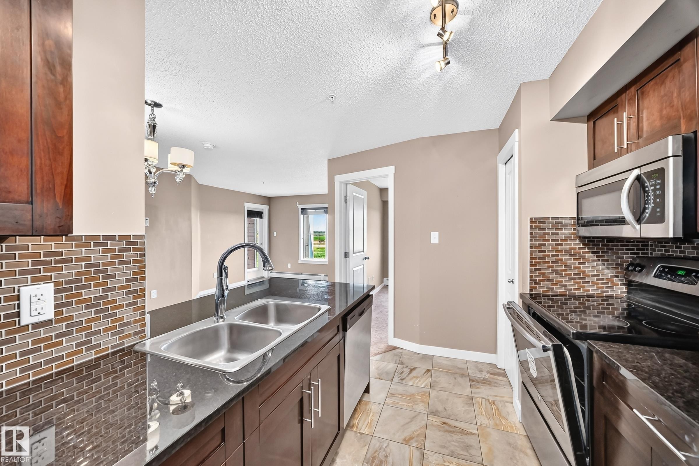 Kitchen featuring dark wood cabinetry, a dual-basin stainless steel sink, black countertops, and a mosaic tile backsplash - 308 18126 77 Street, Edmonton, AB