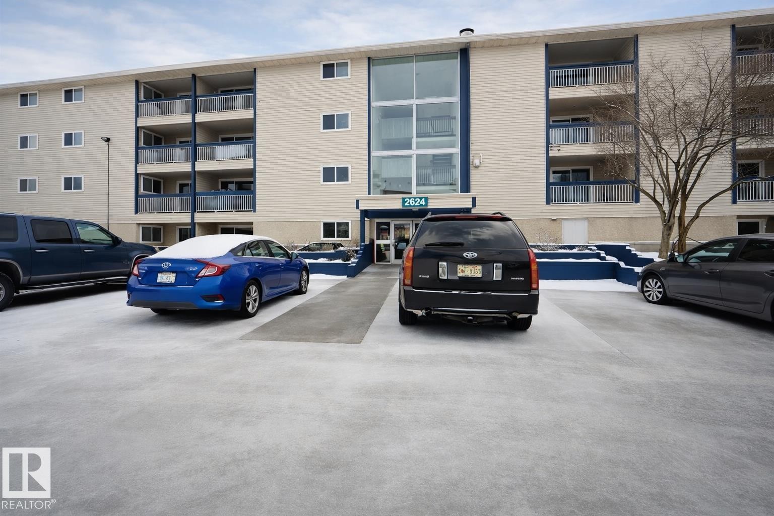 Multi-story building featuring light-colored siding, multiple balconies with dark railings, and a central glass entrance - 107, 2624 Millwoods Road East Nw, Edmonton, AB - Outdoor