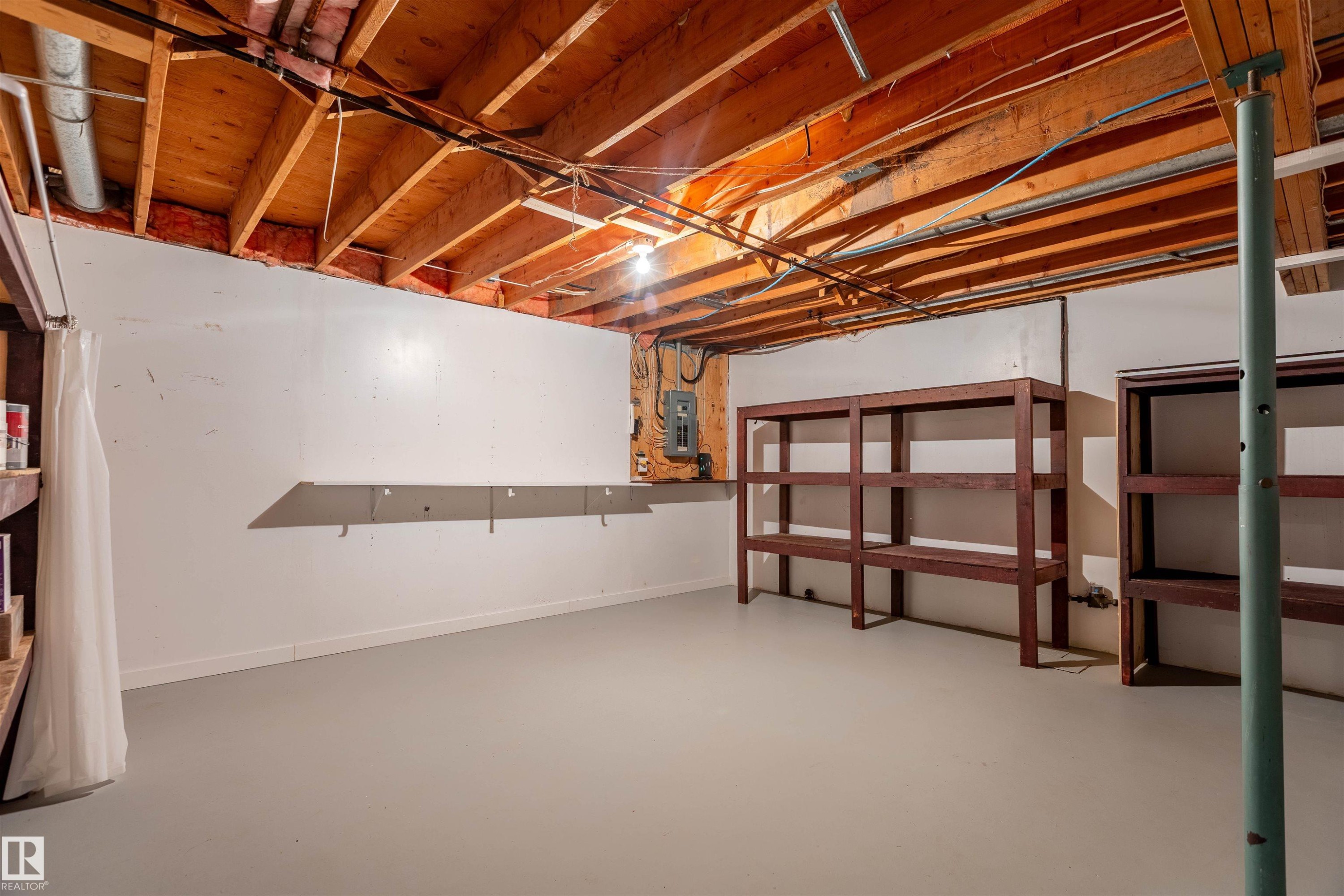 Basement storage space featuring exposed wood joists, painted concrete floor, built-in shelving units, and a visible electrical panel - 3 Glenforest Crescent, Stony Plain, AB