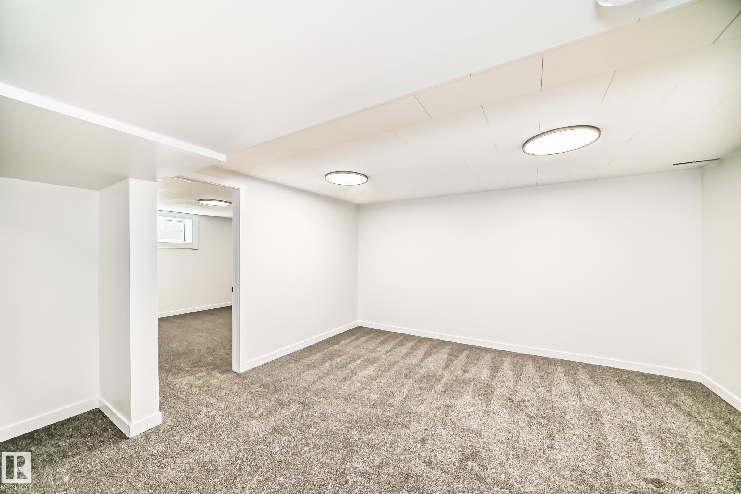 Spacious interior room featuring plush gray carpeting, white painted walls, recessed ceiling lighting, and a visible window opening into an additional space - 4842 116Ave, Edmonton, AB