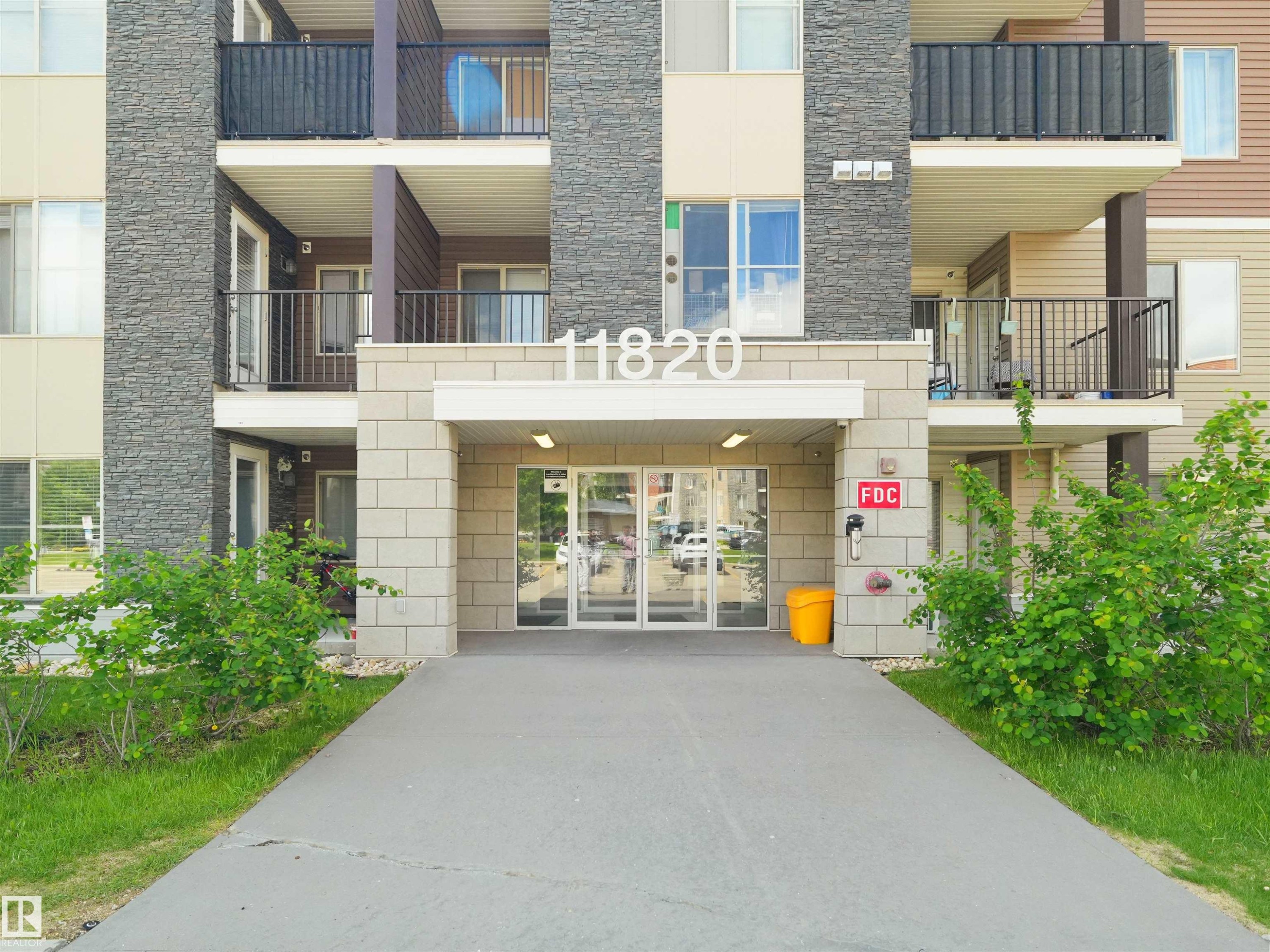 Building exterior featuring a stone facade, multiple balconies with dark railings, a prominent entrance with glass doors, and a concrete pathway - #313 11820 22 Av Sw, Edmonton, AB - Outdoor With Balcony