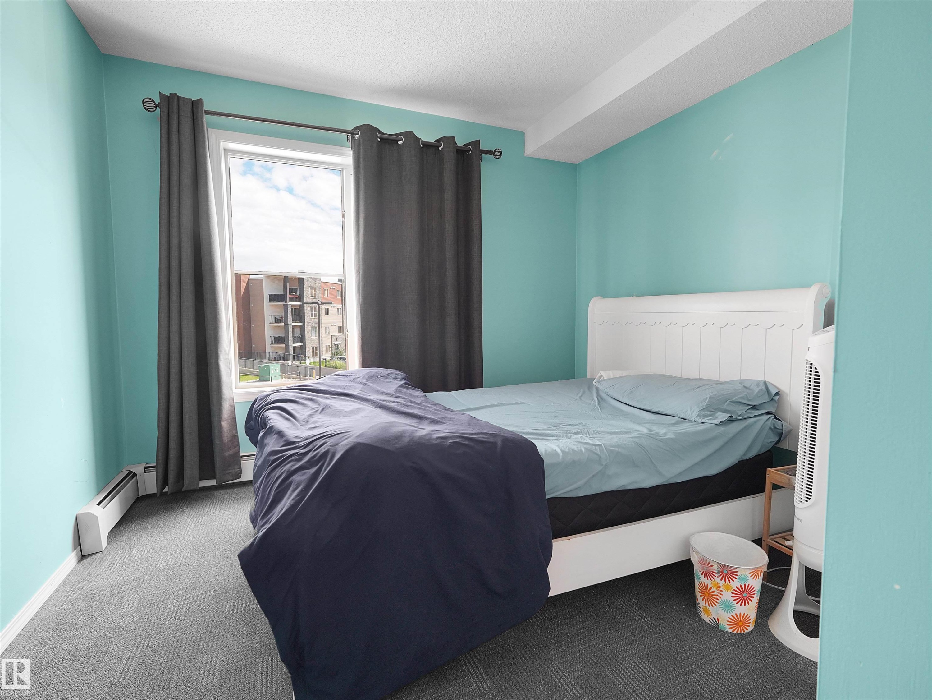 Well-lit interior space featuring a large window, light blue walls, and patterned gray carpet flooring - #313 11820 22 Av Sw, Edmonton, AB - Indoor Photo Showing Bedroom