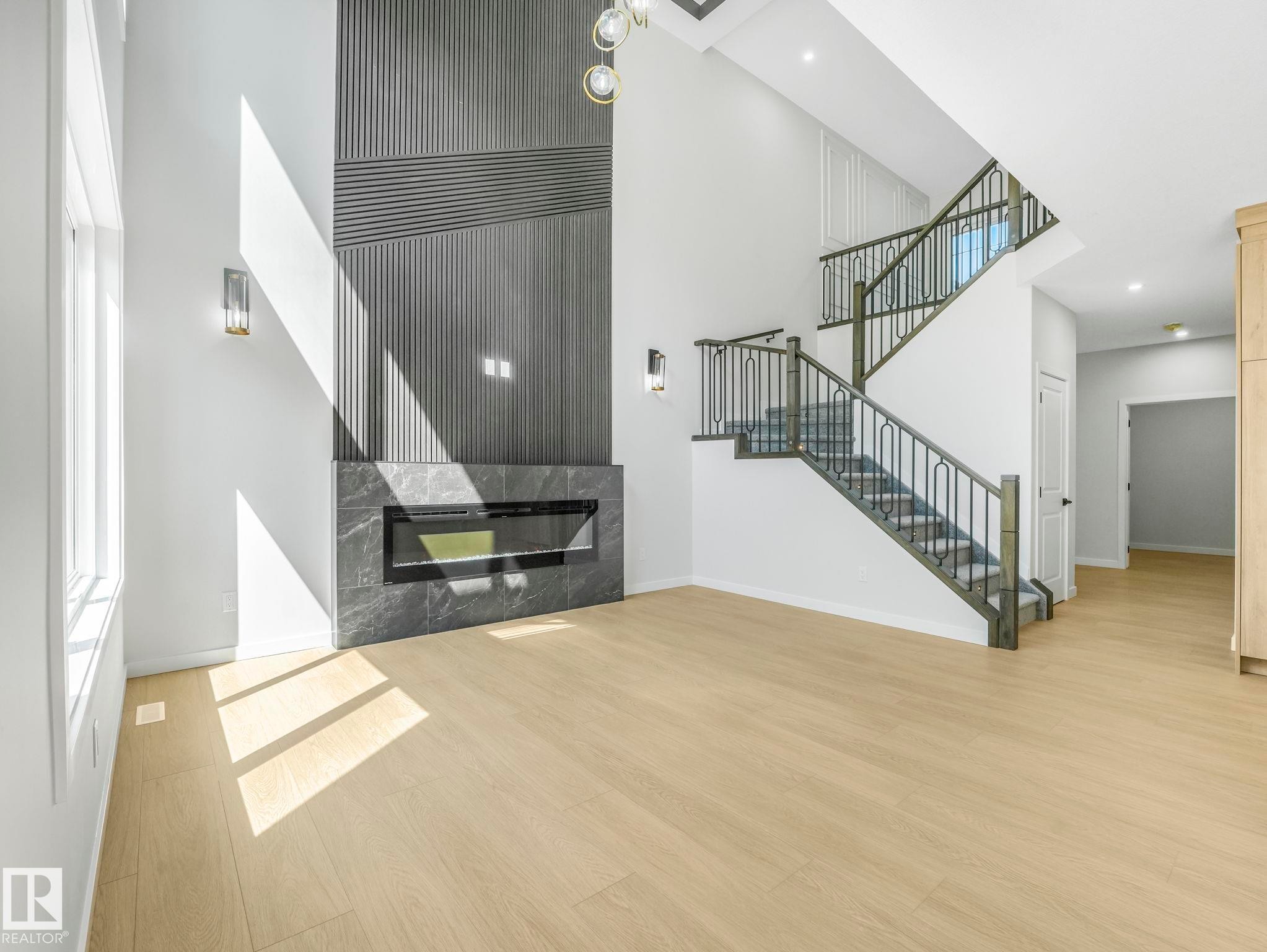Spacious living area featuring a tall fireplace with a linear insert and textured paneling, light wood-finish flooring, a contemporary staircase with dark wood-finish treads and wrought iron balusters, and recessed lighting - 1717 Crossbill Loop, Edmonton, AB - Indoor Photo Showing Other Room With Fireplace