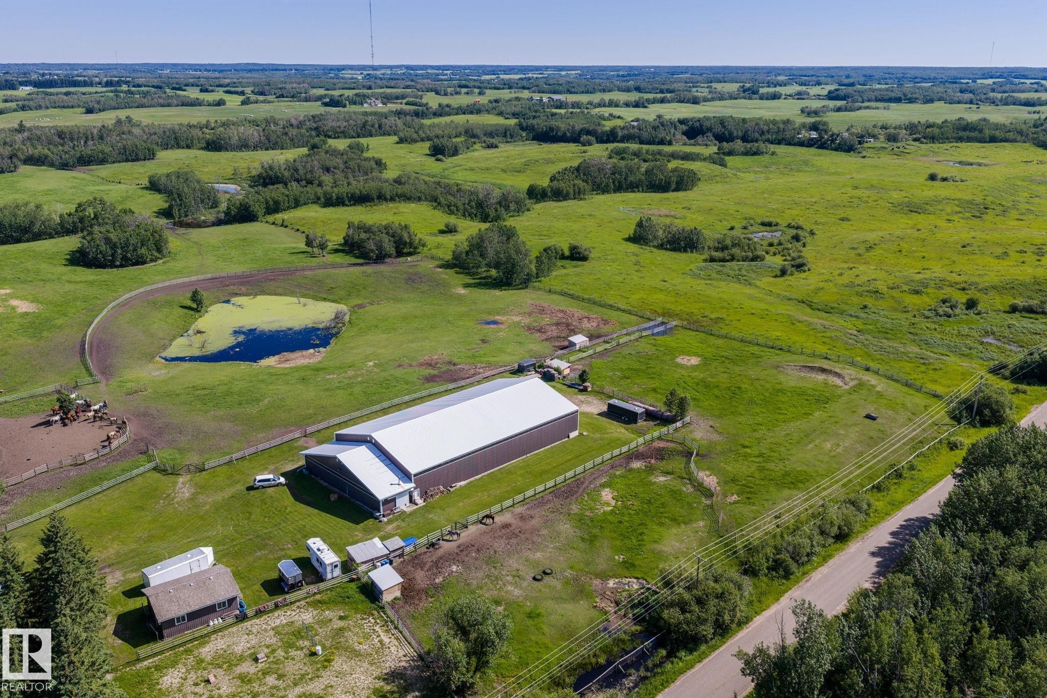 Expansive rural property featuring a large barn structure, a residential building, a visible pond, and fenced paddocks, all surrounded by open green fields and natural tree lines - 860 41 Avenue, Edmonton, AB