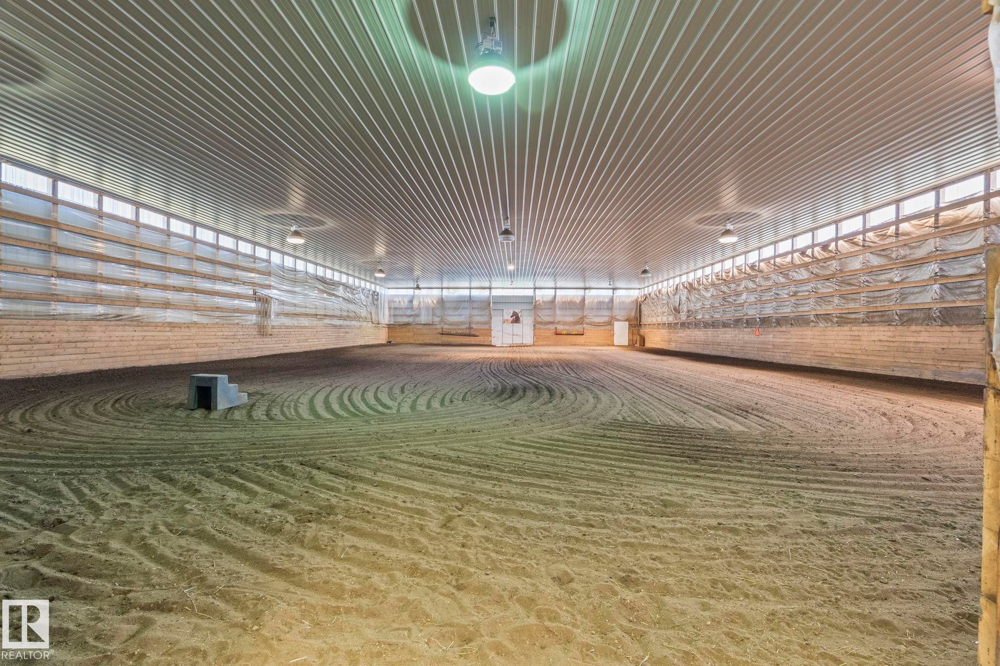 Spacious indoor arena featuring a sand footing, corrugated metal ceiling, perimeter wood paneling, extensive translucent paneling for natural light, and overhead lighting fixtures - 860 41 Avenue, Edmonton, AB