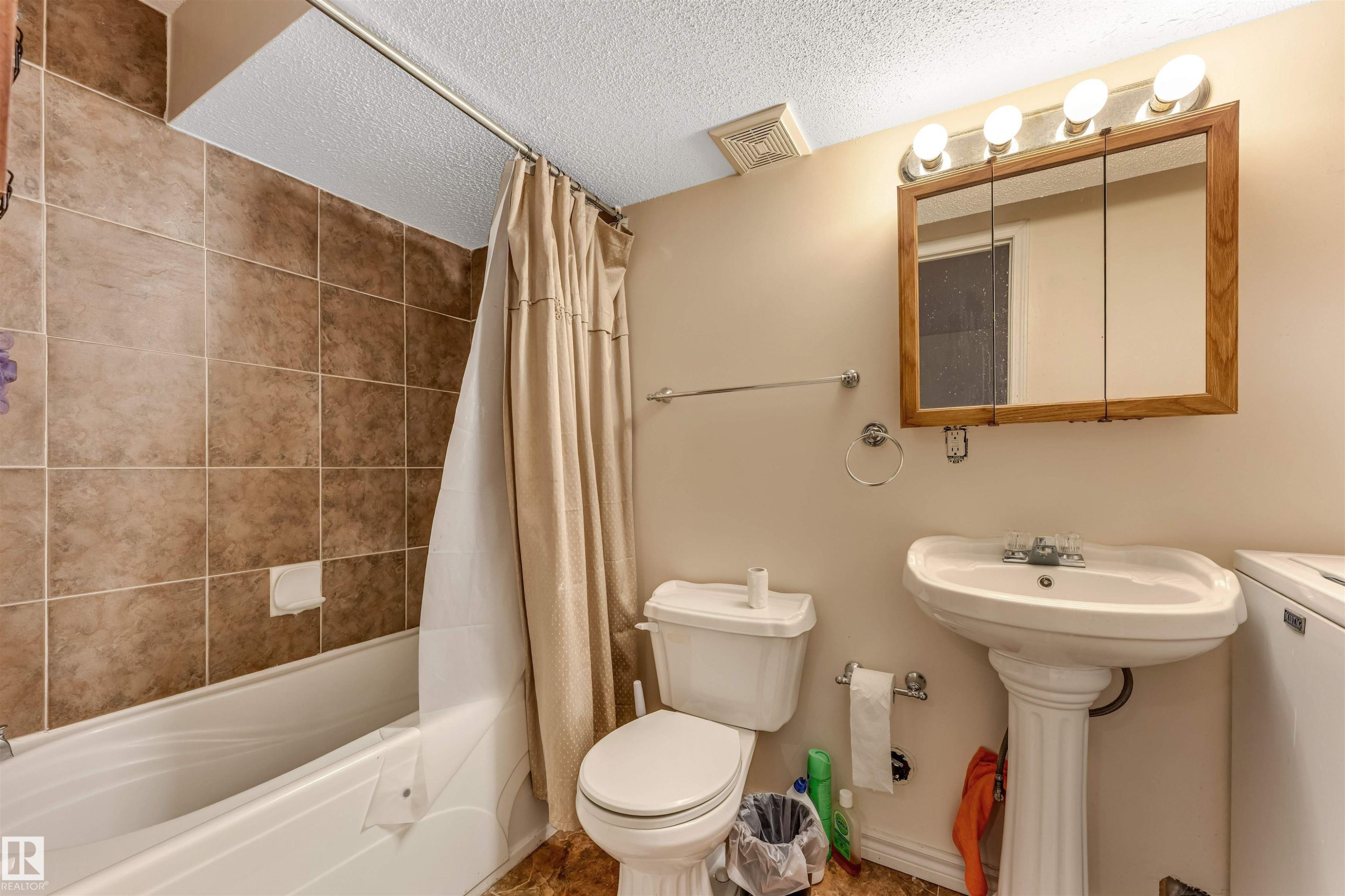 Bathroom featuring a white pedestal sink, a three-door mirrored medicine cabinet with vanity lighting, and a bathtub with full-height brown tiled surround - 3032 31 Avenue, Edmonton, AB - Indoor Photo Showing Bathroom