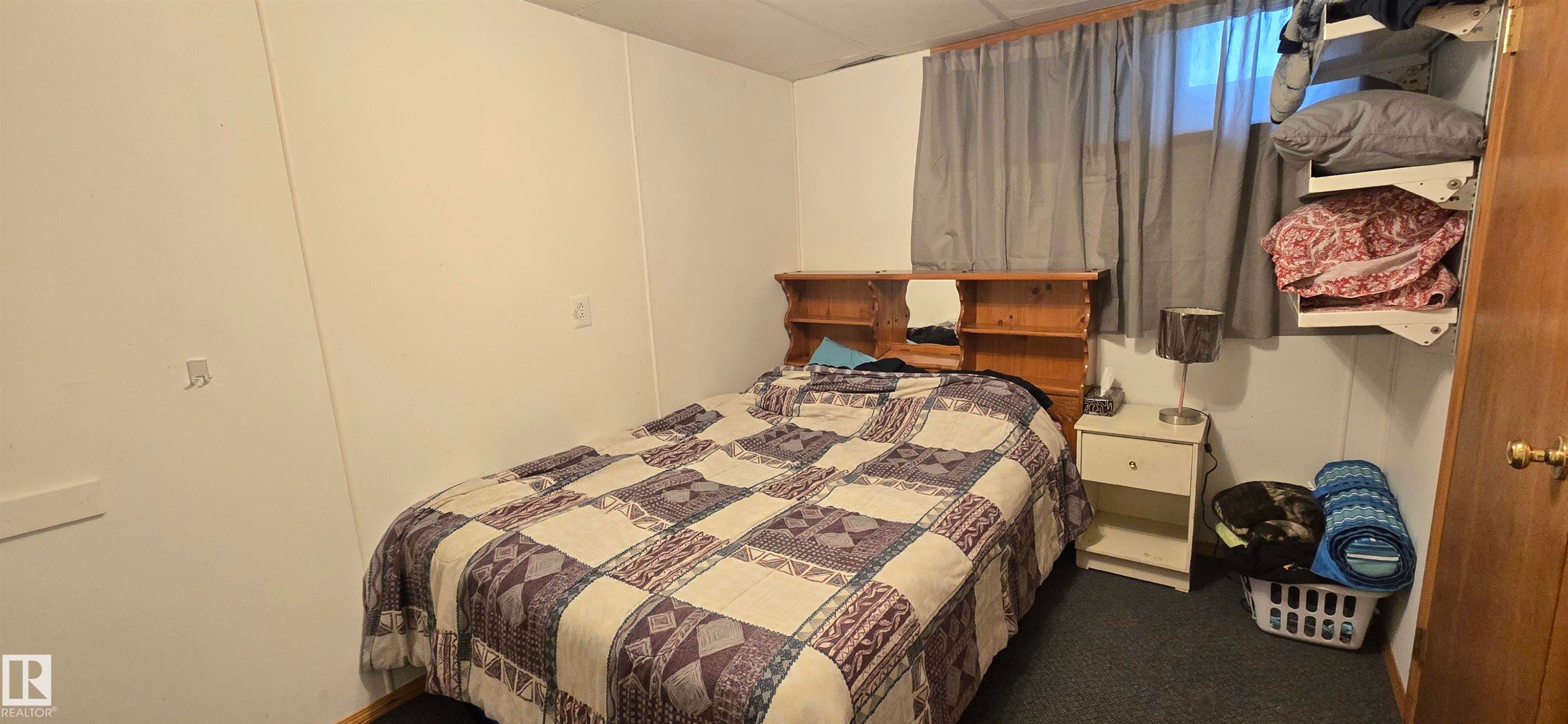 Carpeted room featuring a built-in wooden headboard with shelving and a window with curtains - 5304 49 Street, Two Hills, AB - Indoor Photo Showing Bedroom