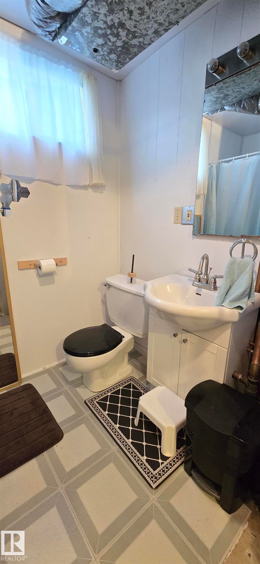 Bathroom featuring light-toned walls, a white sink with a vanity cabinet, a toilet, a mirror, and a window with a sheer curtain - 5304 49 Street, Two Hills, AB - Indoor Photo Showing Bathroom