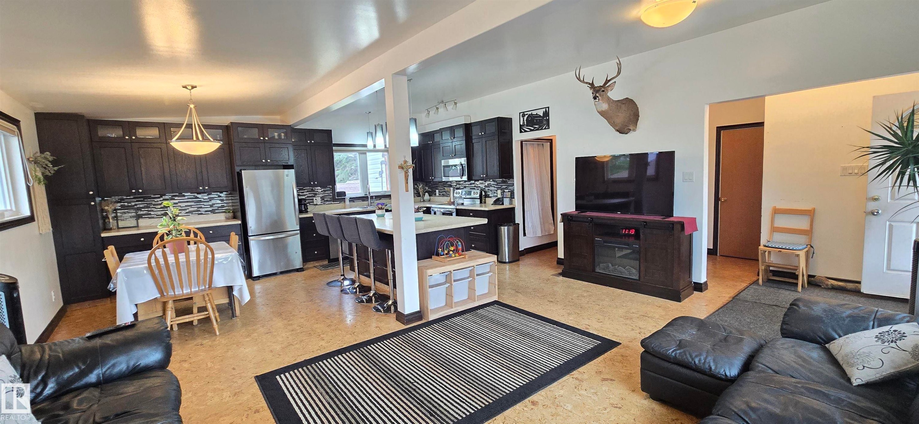 Open-concept living space featuring wood-finish flooring, a contemporary kitchen with dark cabinetry and stainless steel appliances, and a breakfast bar with pendant lighting - 5304 49 Street, Two Hills, AB - Indoor