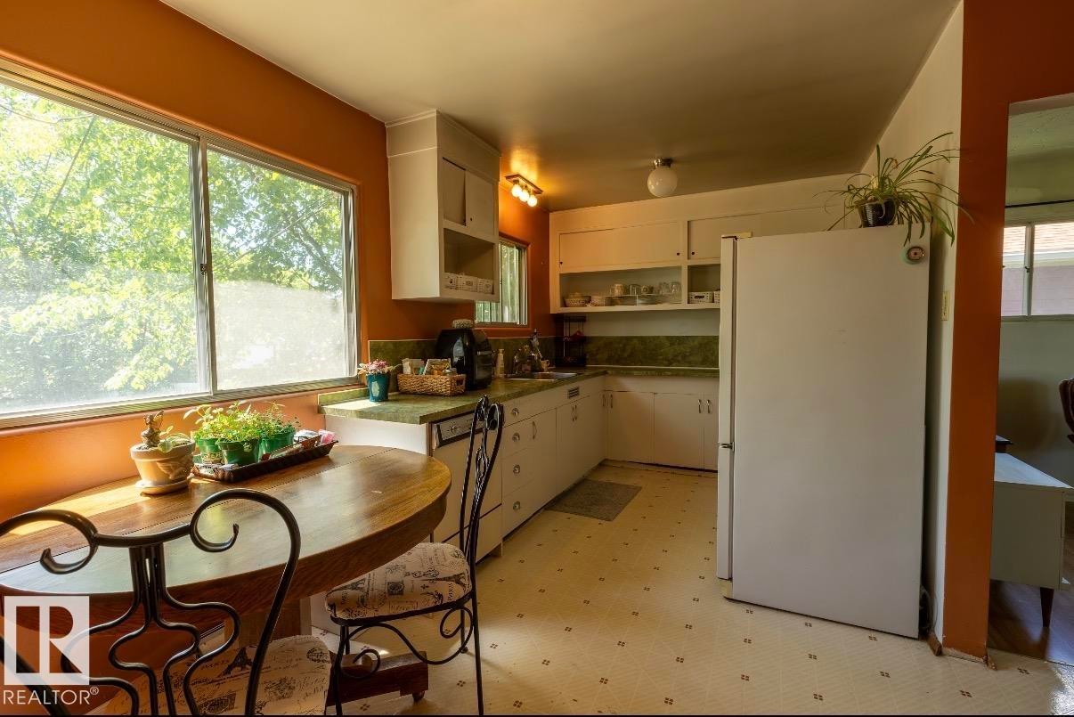 Kitchen featuring a built-in wood-finish dining table, white cabinetry, integrated dishwasher, and a large window overlooking lush greenery - 8719 67 Avenue Nw, Edmonton, AB - Indoor