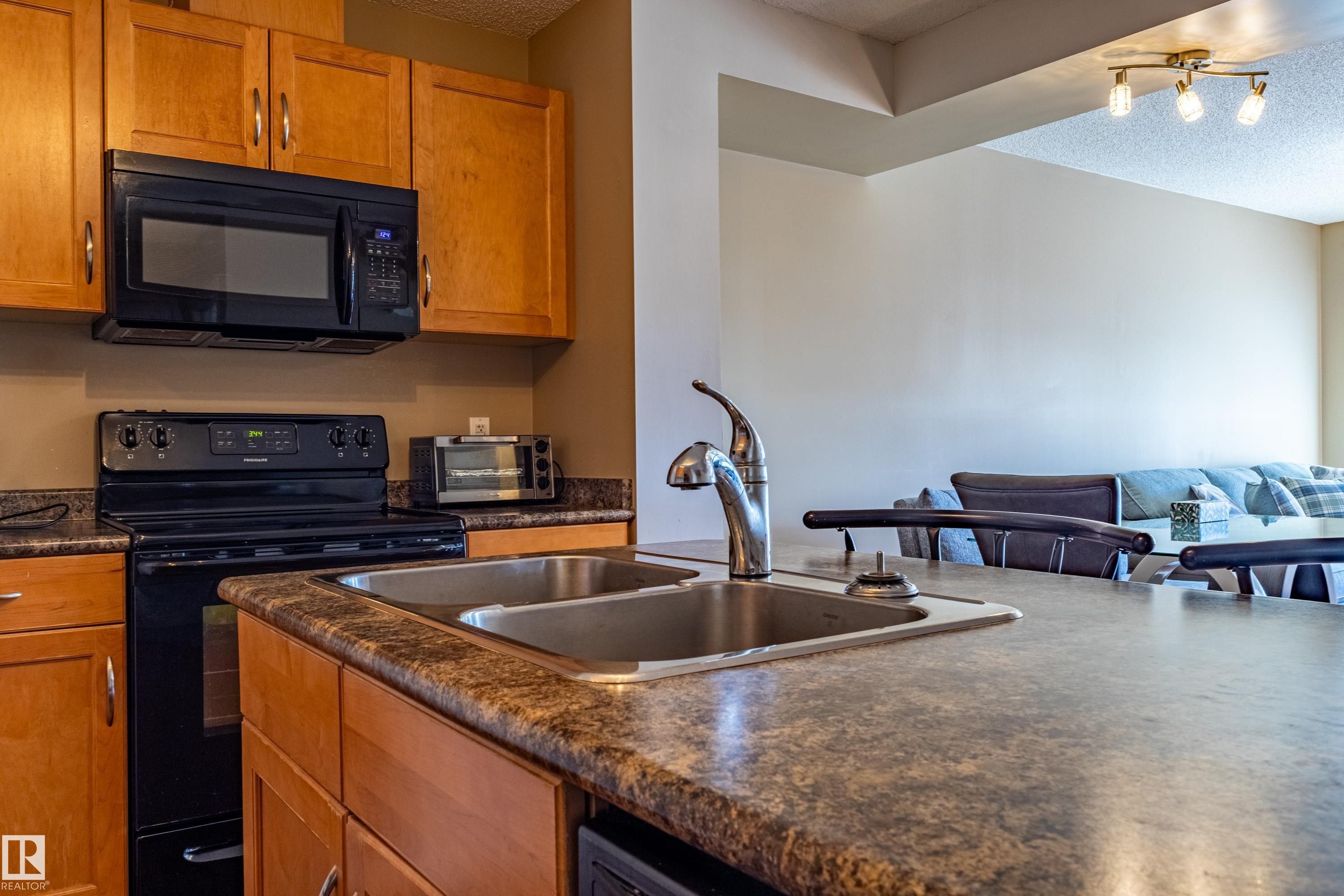Kitchen featuring a double basin sink with a gooseneck faucet, light wood cabinetry, and a black range with an overhead microwave - 13 1816 Rutherford Road, Edmonton, AB