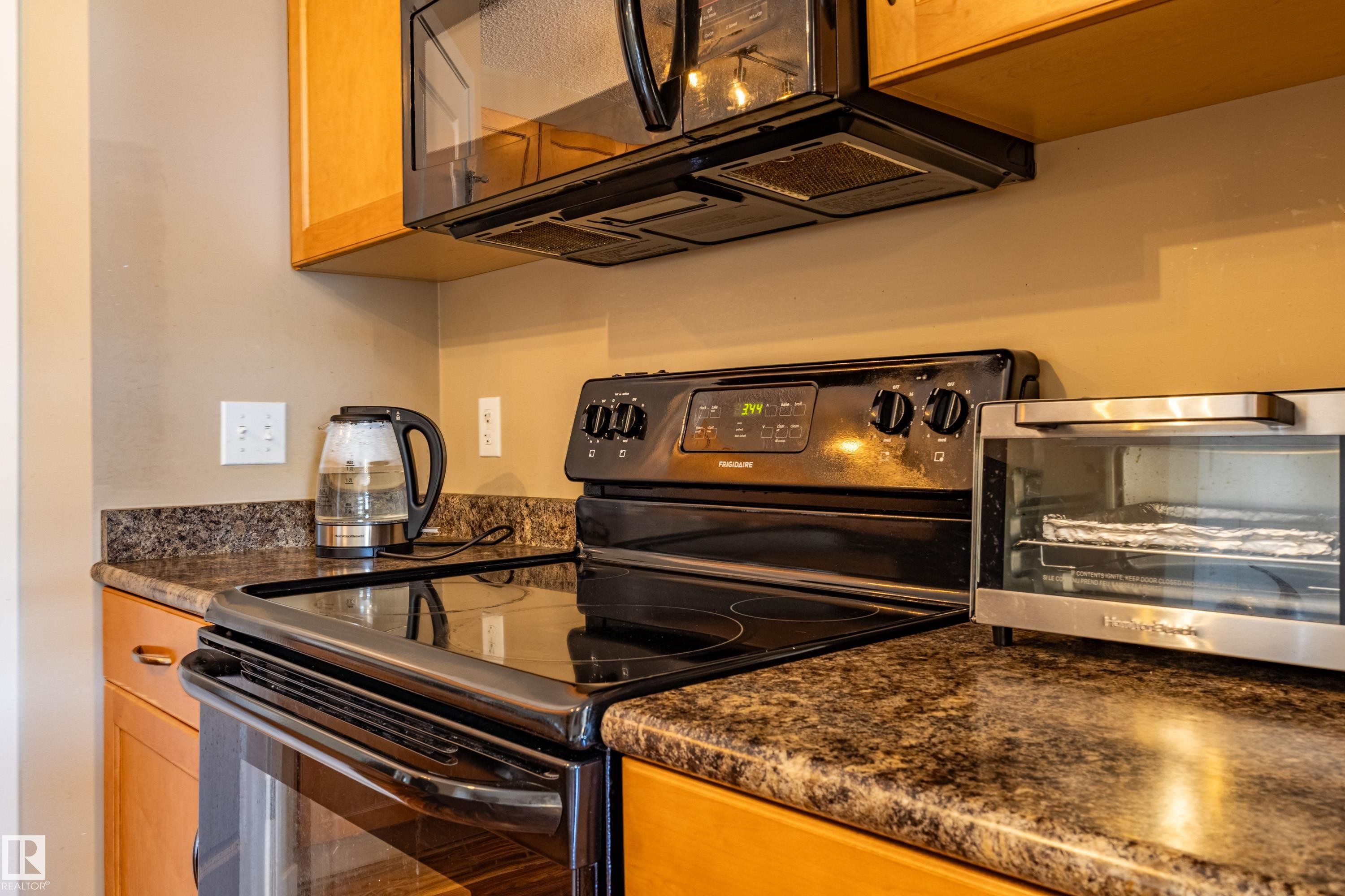 Kitchen featuring a black electric range with an overhead microwave, dark speckled countertops, and light wood cabinetry - 13 1816 Rutherford Road, Edmonton, AB