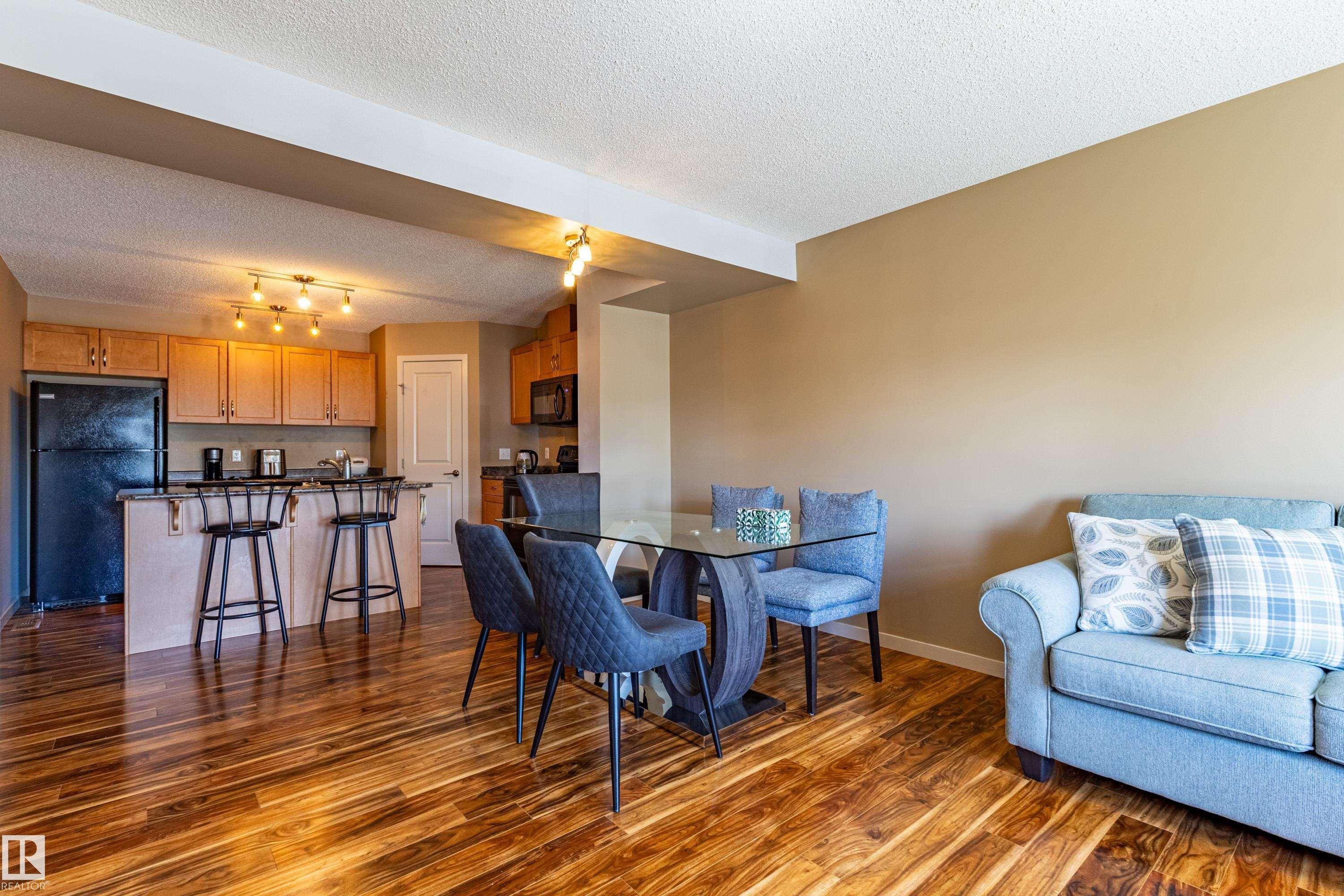 Open-concept living area featuring rich wood-finish flooring, a kitchen with black appliances and light wood cabinetry, and an eat-at island with a dark countertop - 13 1816 Rutherford Road, Edmonton, AB