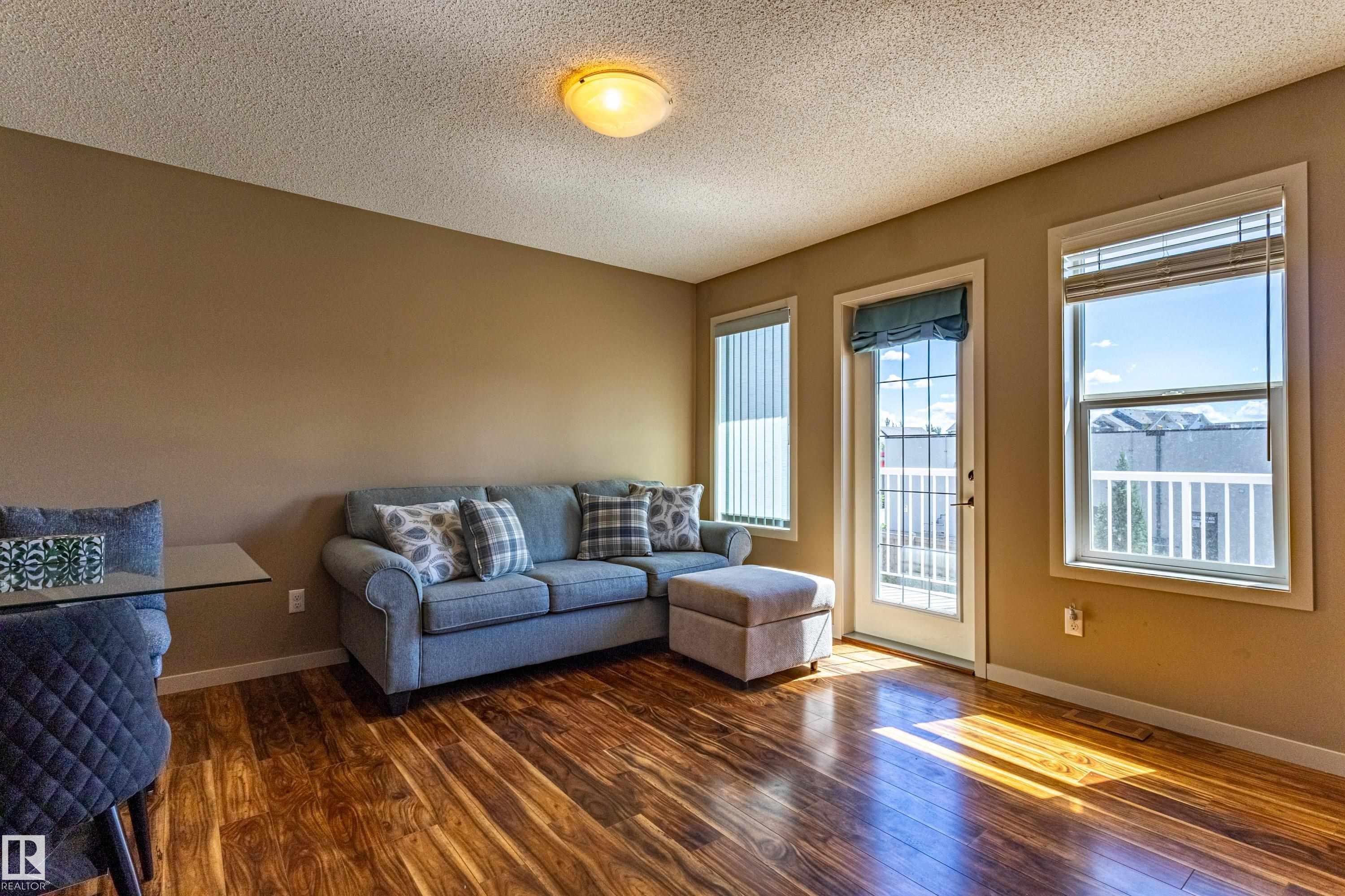 Living area featuring rich wood-finish flooring, neutral-toned walls, and multiple windows with white blinds - 13 1816 Rutherford Road, Edmonton, AB