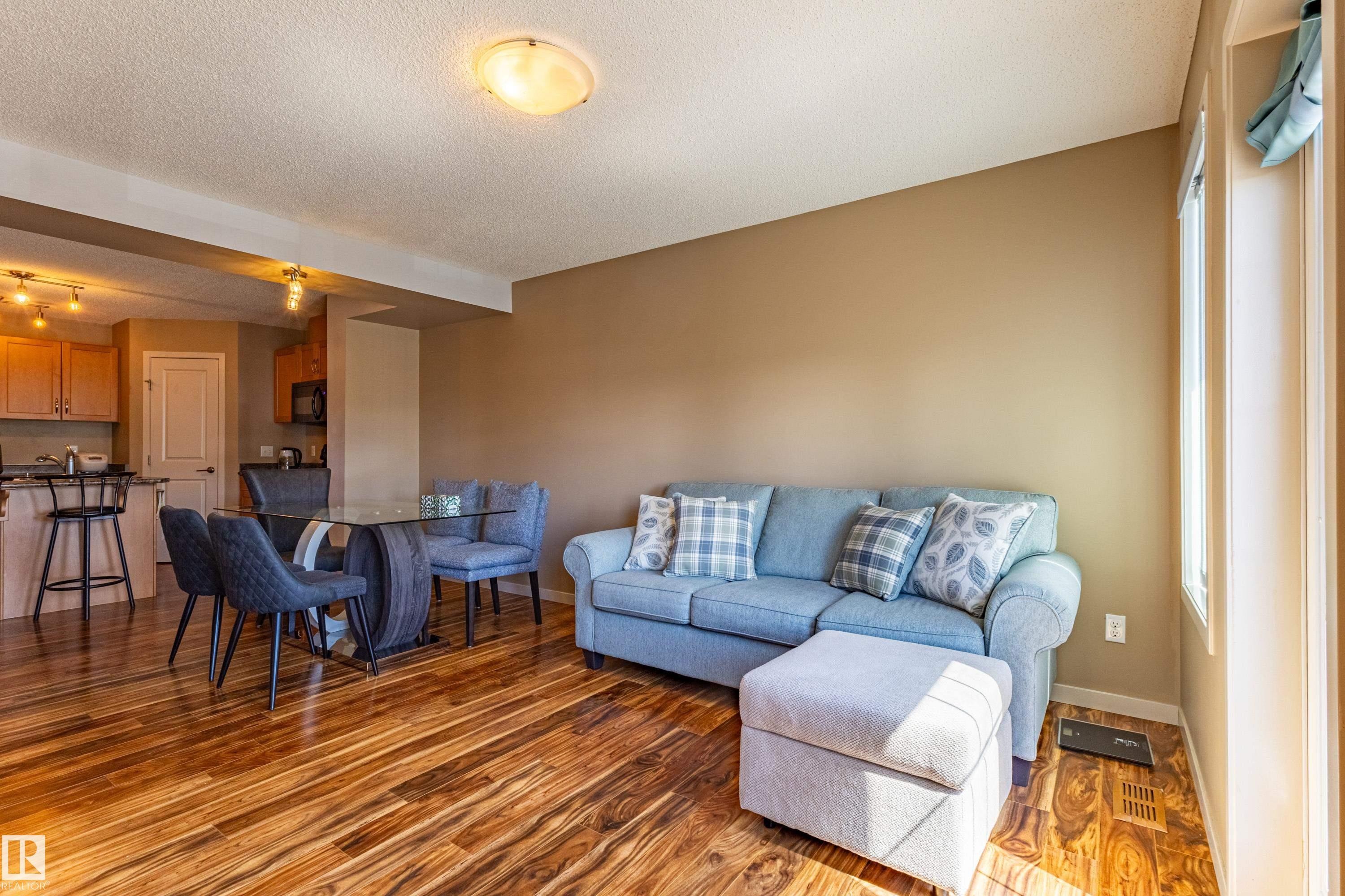 Open-concept living area featuring rich wood-finish flooring, a contemporary glass-top dining table, and direct sightlines to the kitchen - 13 1816 Rutherford Road, Edmonton, AB