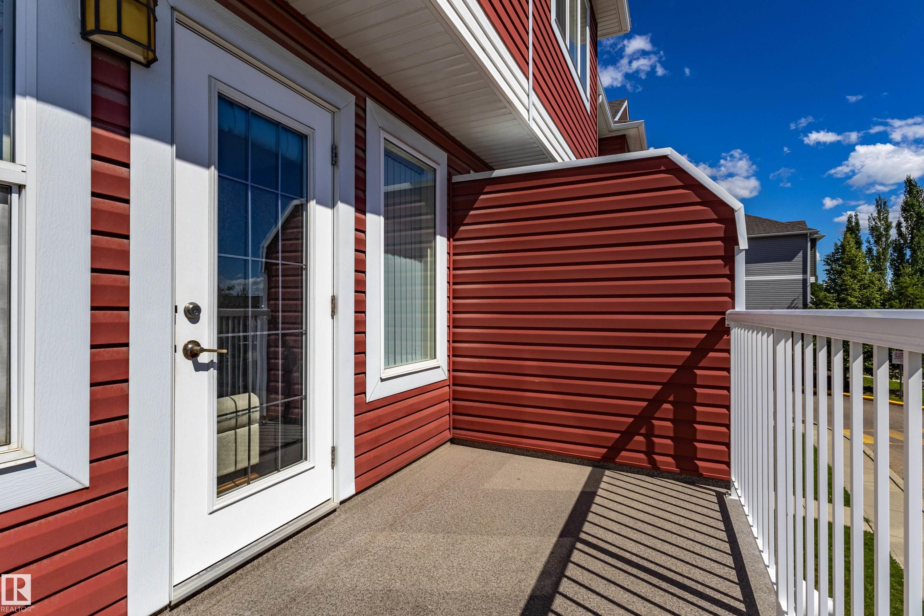 Private balcony featuring a glass-paneled door, two fixed windows, white railing, and red siding - 13 1816 Rutherford Road, Edmonton, AB