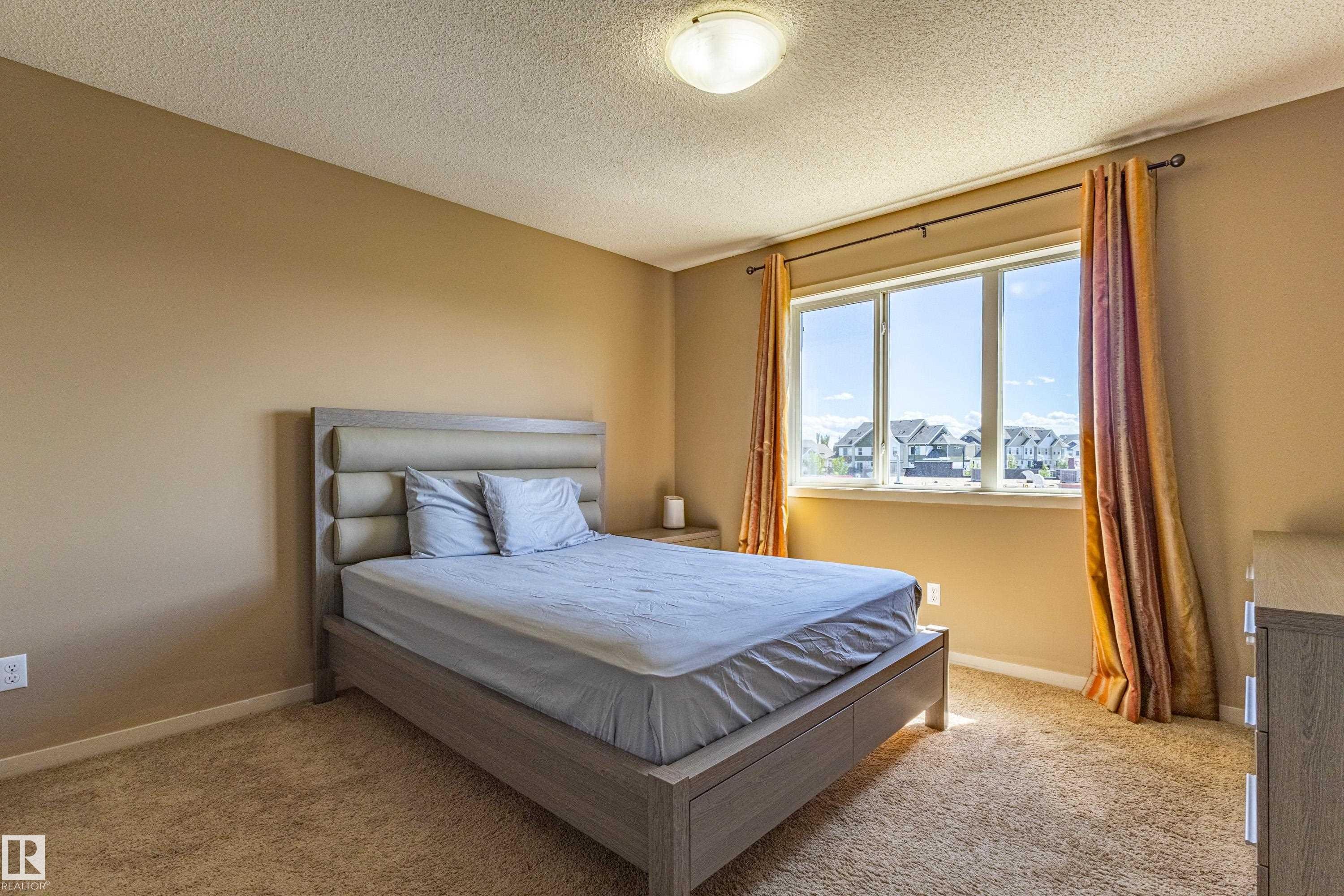 Carpeted room featuring a large window with exterior views, warm neutral wall tones, and a ceiling-mounted light fixture - 13 1816 Rutherford Road, Edmonton, AB