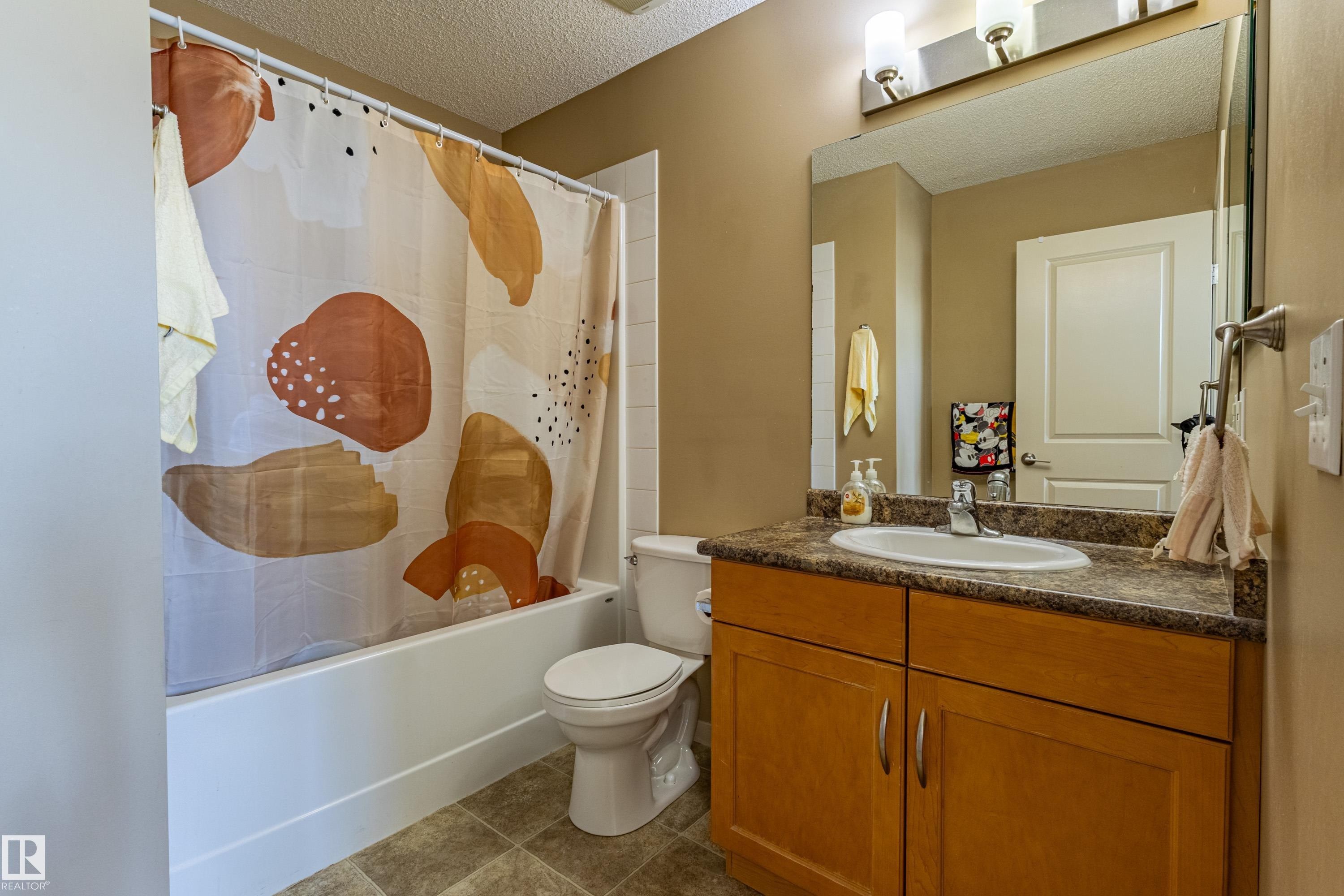 Bathroom featuring a wood-finish vanity with a built-in sink, a large wall-mounted mirror, and a full-size bathtub with white tile surround - 13 1816 Rutherford Road, Edmonton, AB