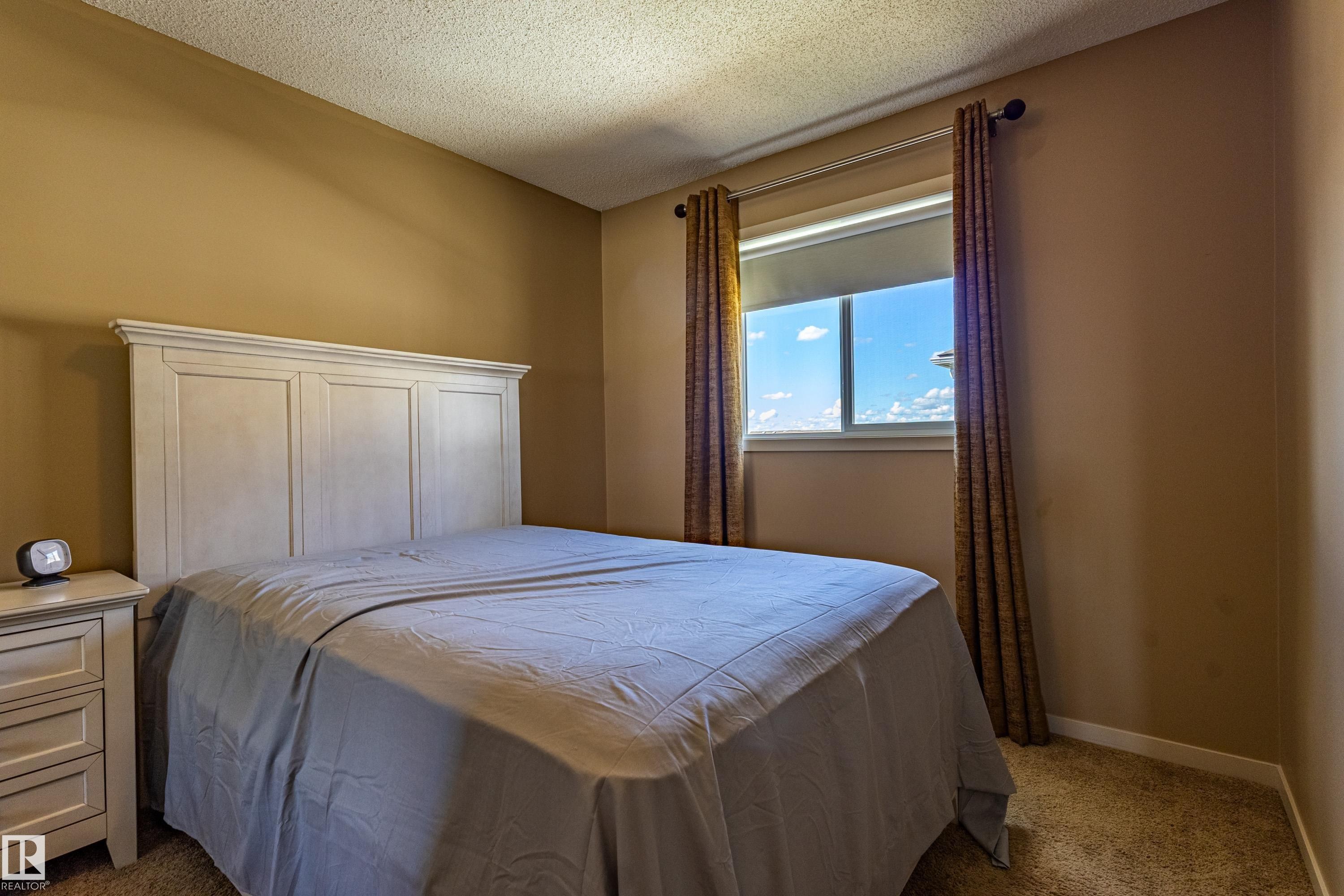 Window with roller shade and decorative drapery panels, complemented by an off-white headboard and matching nightstand - 13 1816 Rutherford Road, Edmonton, AB