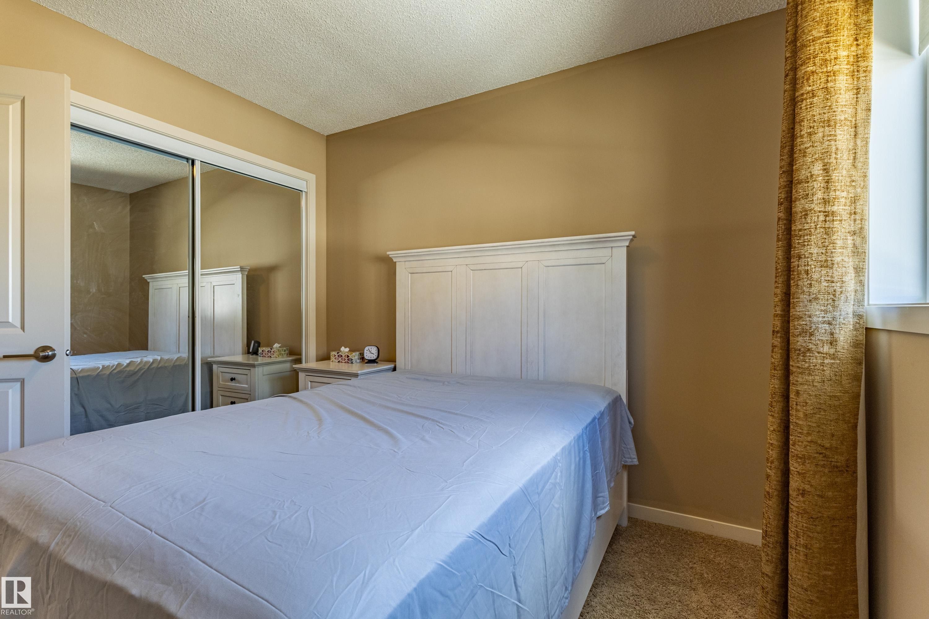 Bedroom featuring beige wall paint, light-colored carpeting, a white paneled headboard, and a window with a textured curtain - 13 1816 Rutherford Road, Edmonton, AB