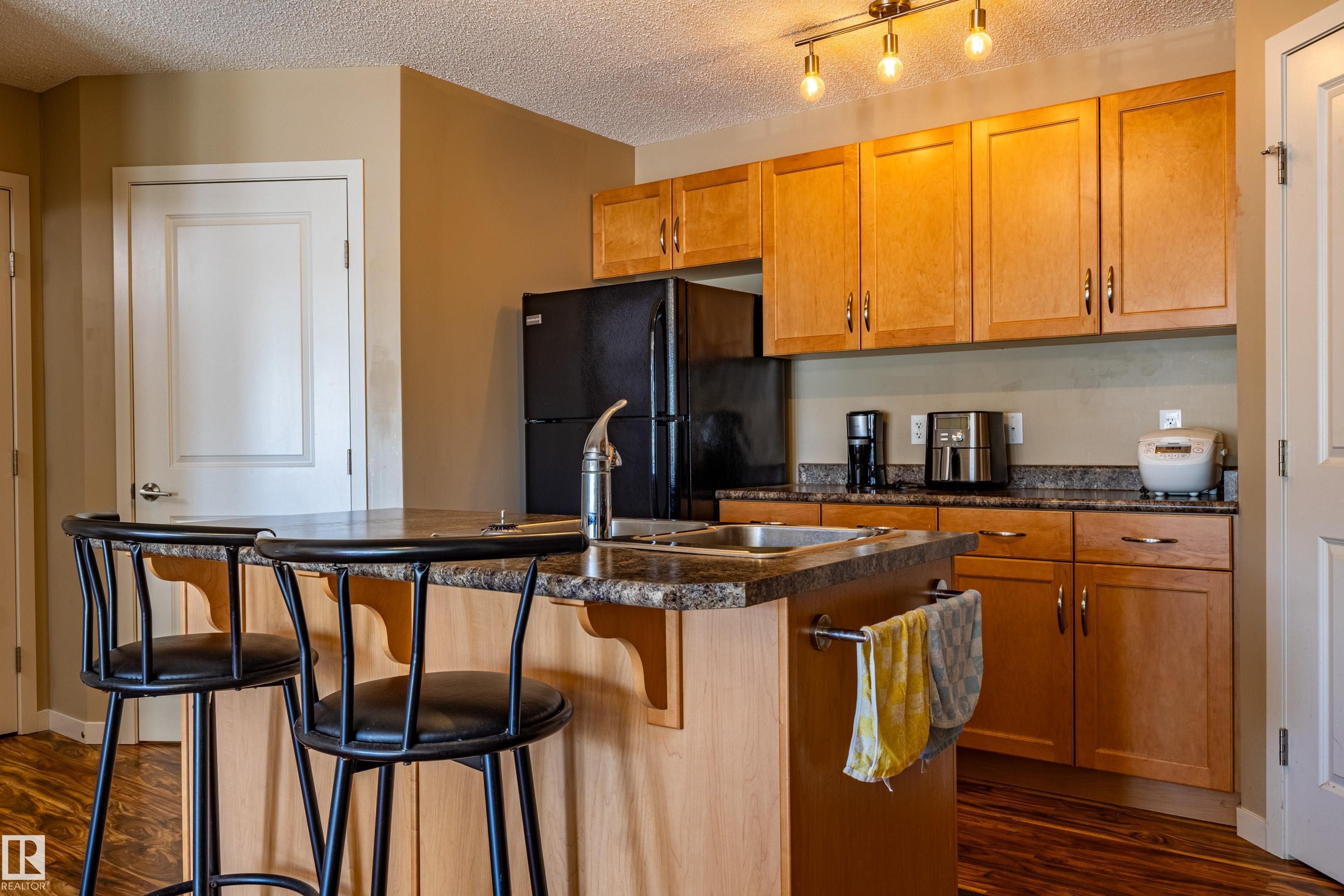 Kitchen featuring wood-finish cabinetry, dark countertop surfaces, a black refrigerator, and an island with an integrated sink - 13 1816 Rutherford Road, Edmonton, AB