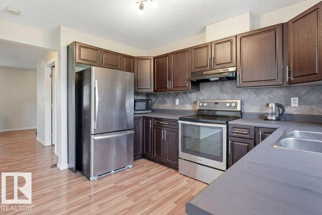 Kitchen featuring wood-finish flooring, dark wood cabinetry, stainless steel appliances, and a tiled backsplash - 30 1404 Hermitage Road, Edmonton, AB