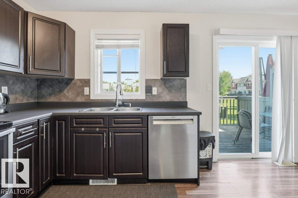 Kitchen featuring dark wood cabinetry, a stainless steel dishwasher, a double basin sink, and a tiled backsplash - 30 1404 Hermitage Road, Edmonton, AB