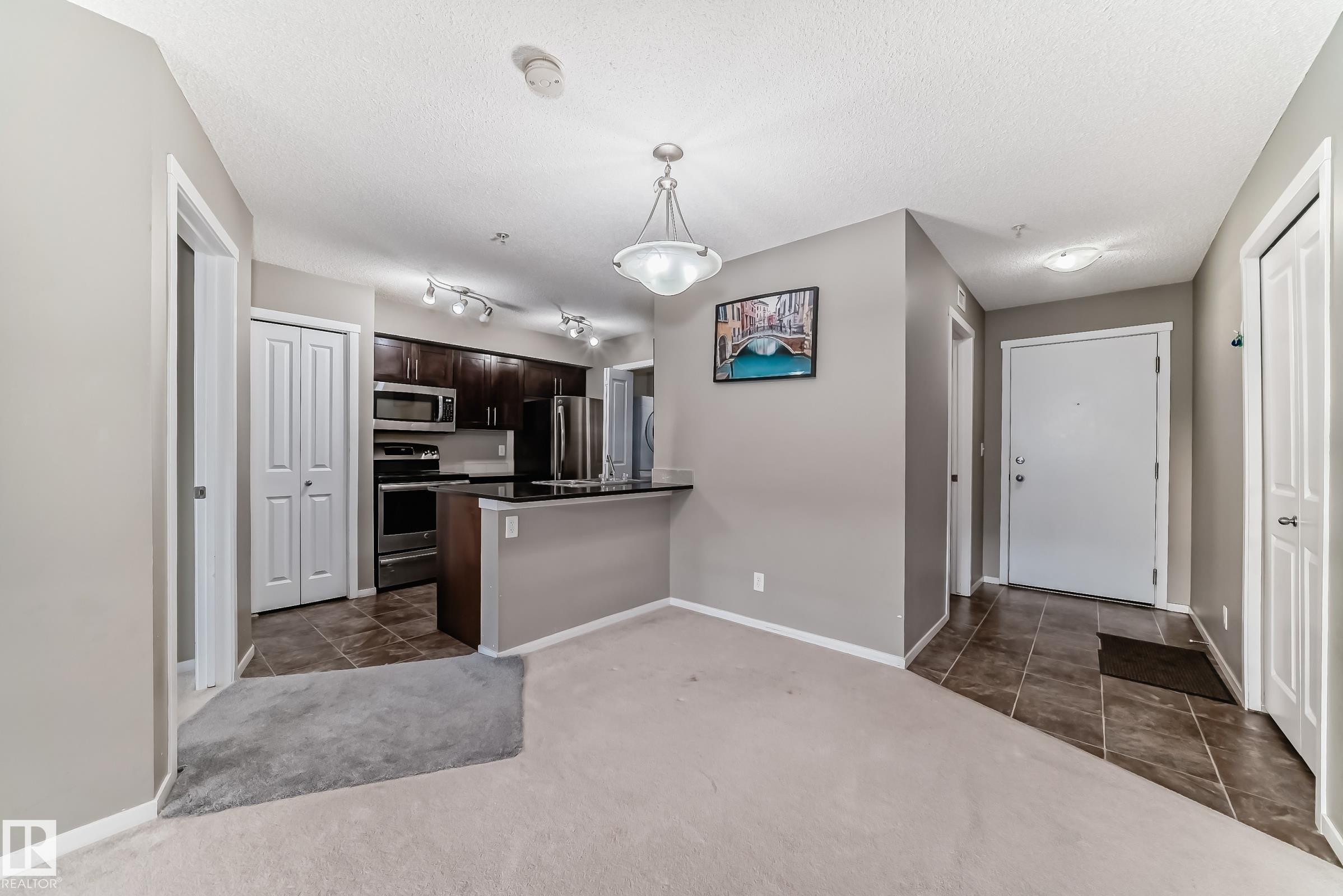 Open-concept living space featuring a kitchen with dark wood-finish cabinetry, stainless steel appliances, and a black countertop with a breakfast bar - #121 111 Watt Cm Sw, Edmonton, AB - Indoor Photo Showing Kitchen