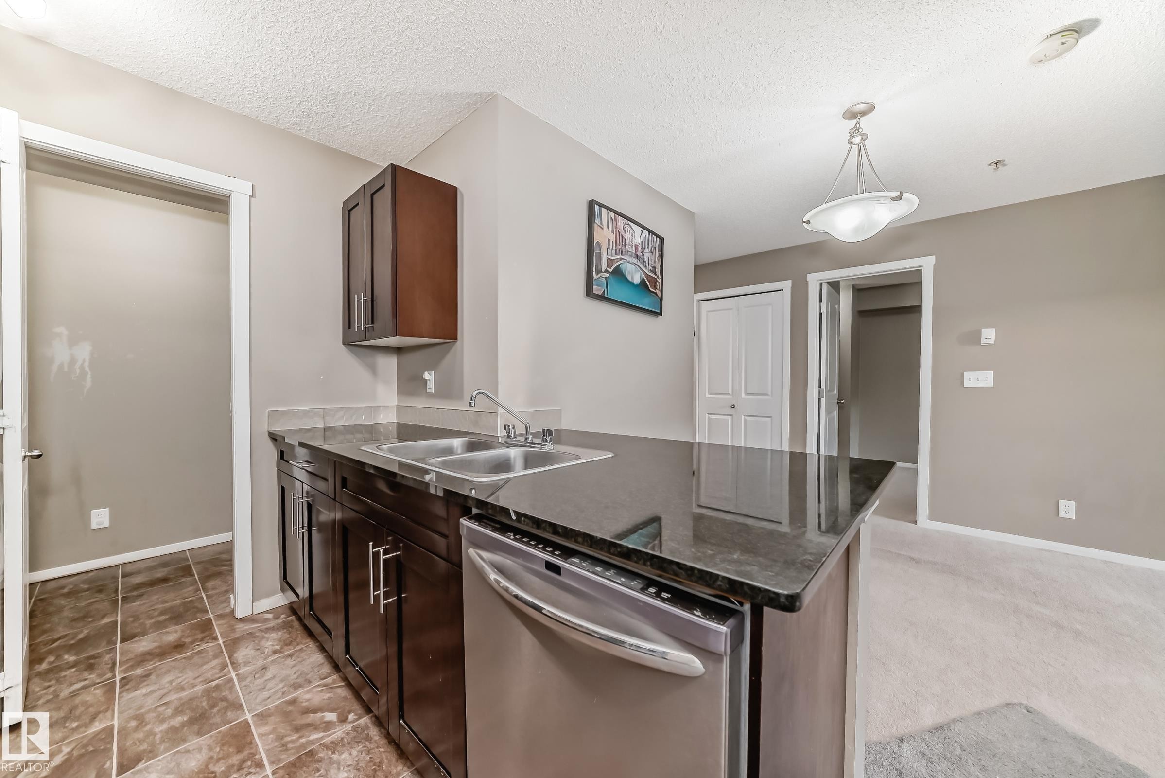 Kitchen island featuring a double basin stainless steel sink, dark cabinetry, a built-in dishwasher with a stainless steel finish, and a dark stone countertop - #121 111 Watt Cm Sw, Edmonton, AB - Indoor Photo Showing Kitchen With Double Sink