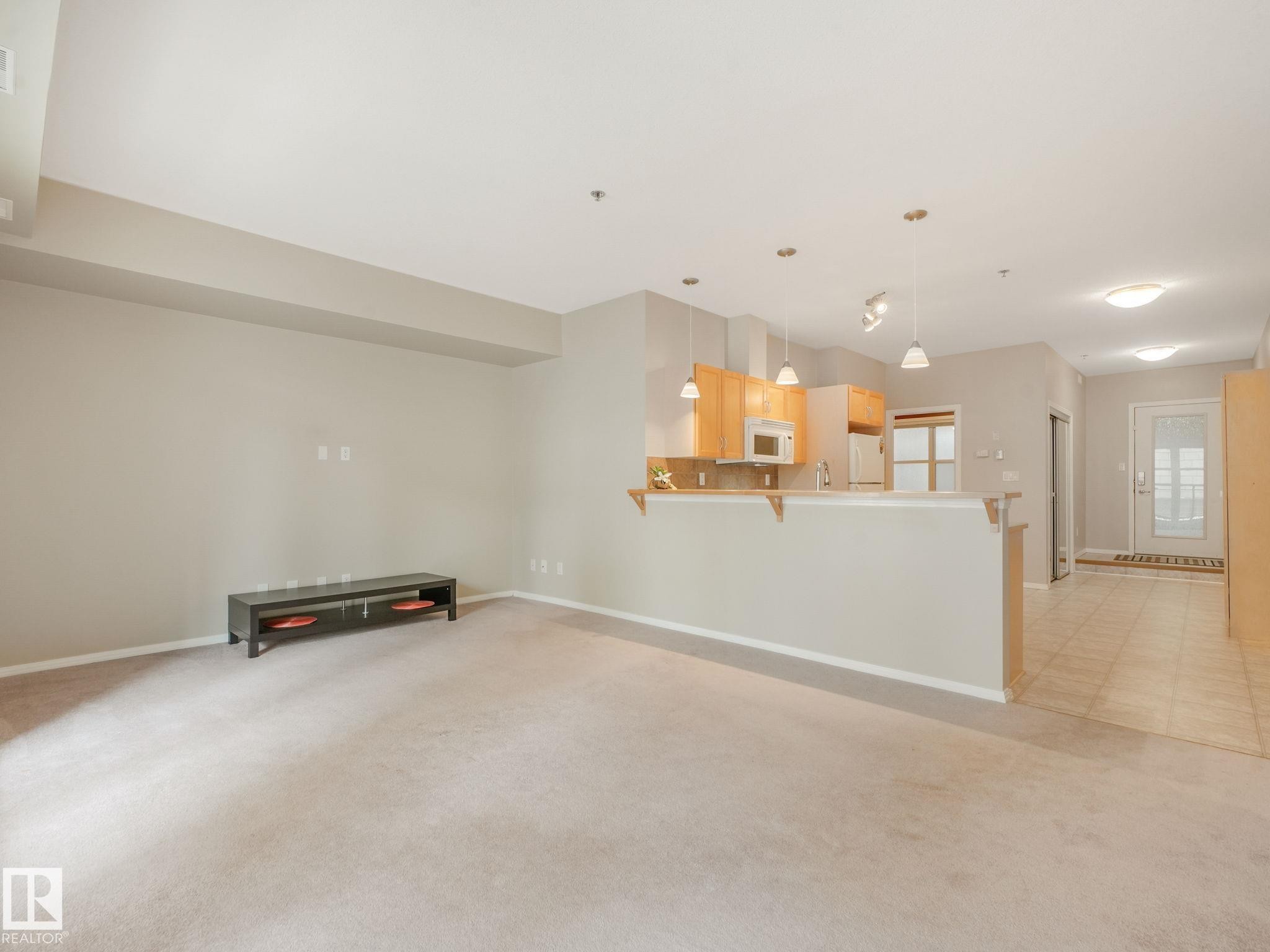 Open-concept living space featuring neutral carpet, recessed lighting, a breakfast bar, light wood-finish cabinetry, and a white refrigerator - 150 4827 104A Street, Edmonton, AB