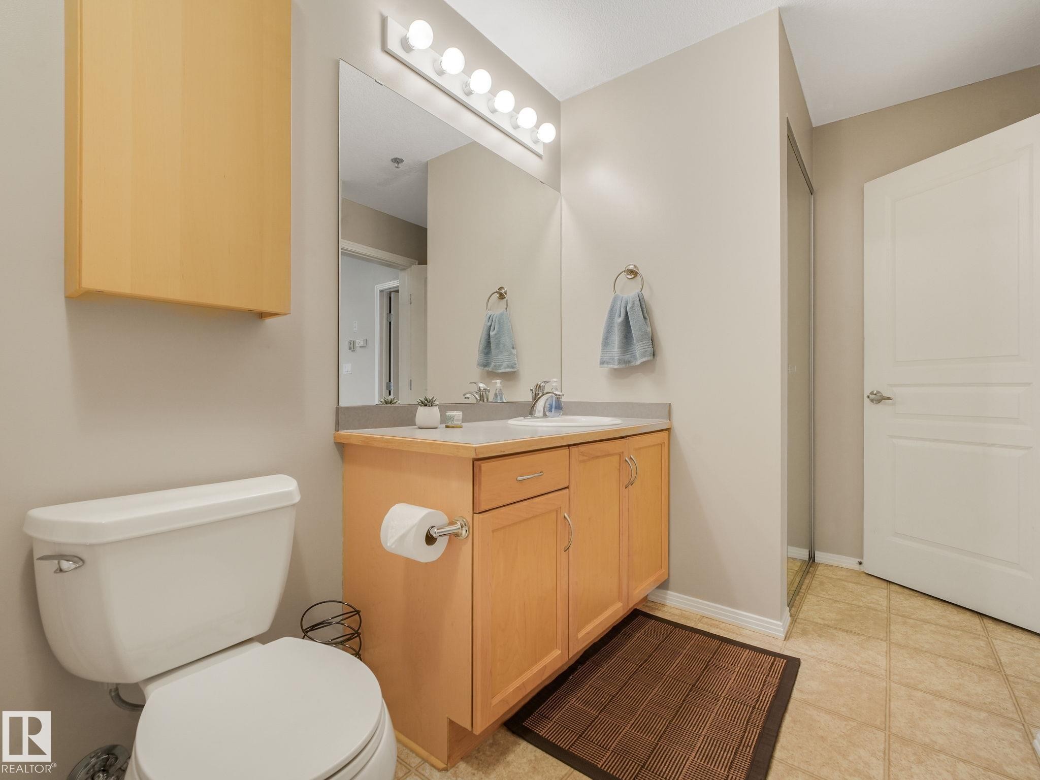Bathroom featuring a light wood-finish vanity with a light countertop, a rectangular mirror with vanity lighting, a wall-mounted storage cabinet, and tiled flooring - 150 4827 104A Street, Edmonton, AB